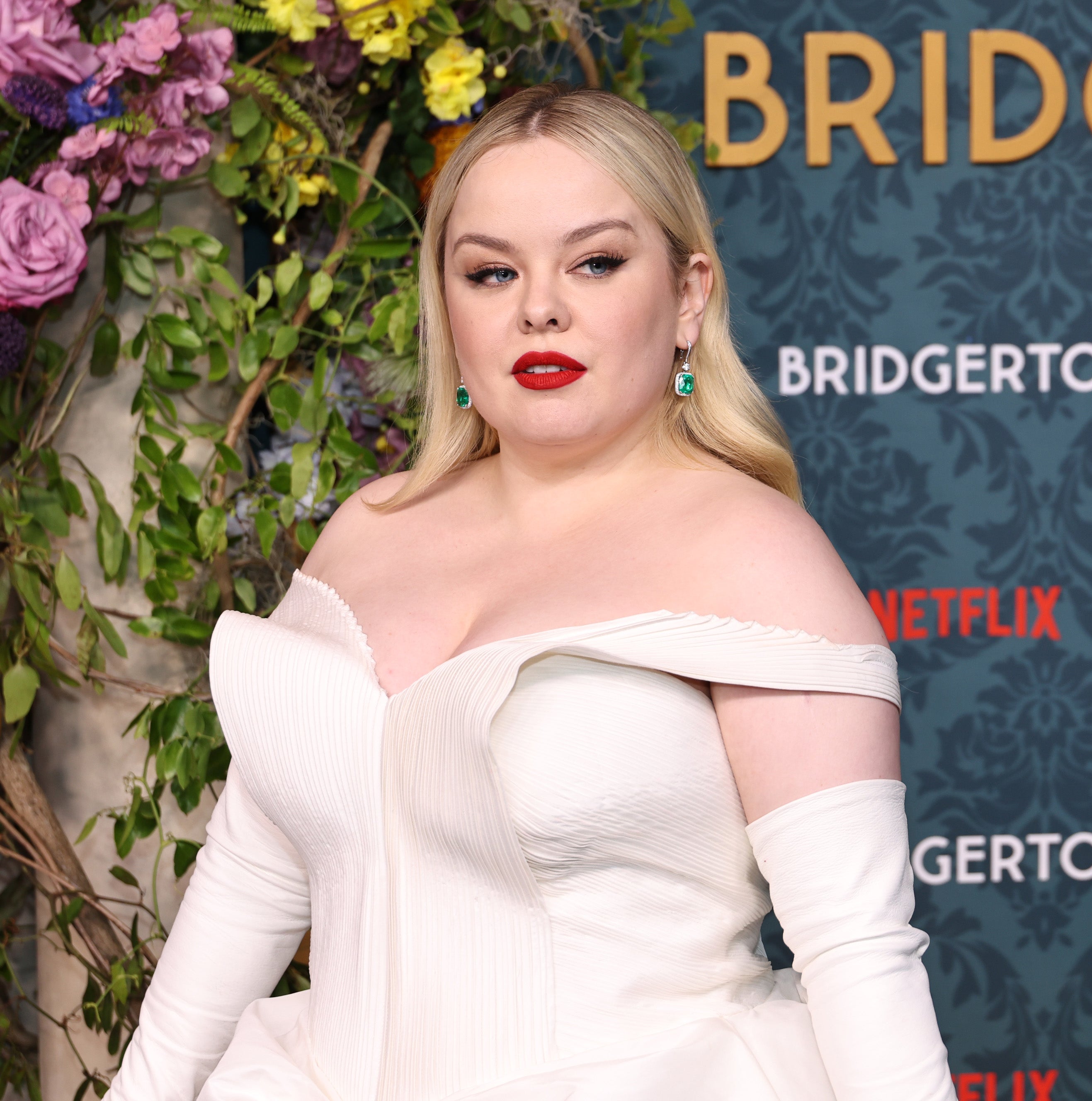 Nicola Coughlan at the "Bridgerton" event, wearing an off-the-shoulder gown, standing in front of a floral and themed backdrop