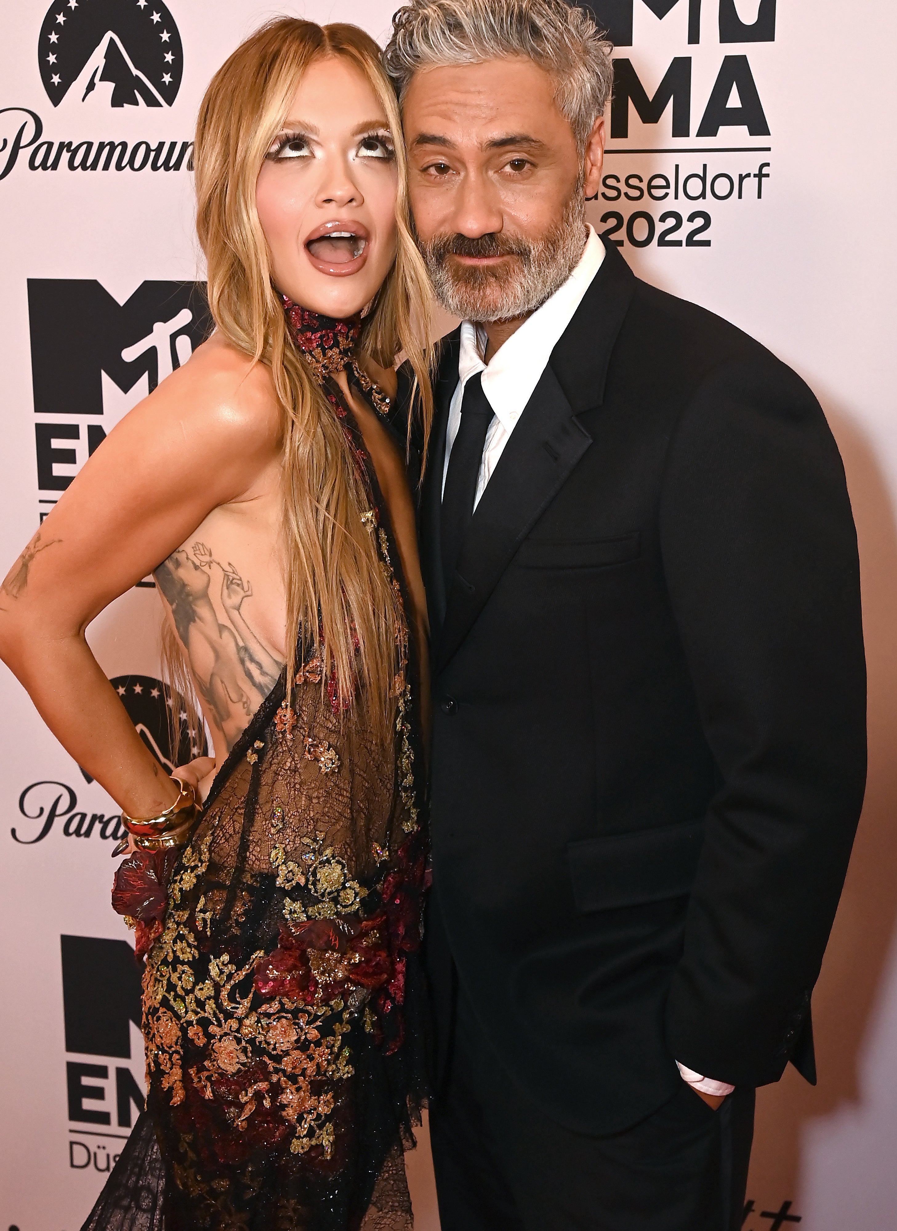 Rita Ora in a patterned dress poses with Taika Waititi in a suit at the MTV EMAs 2022 red carpet in Düsseldorf