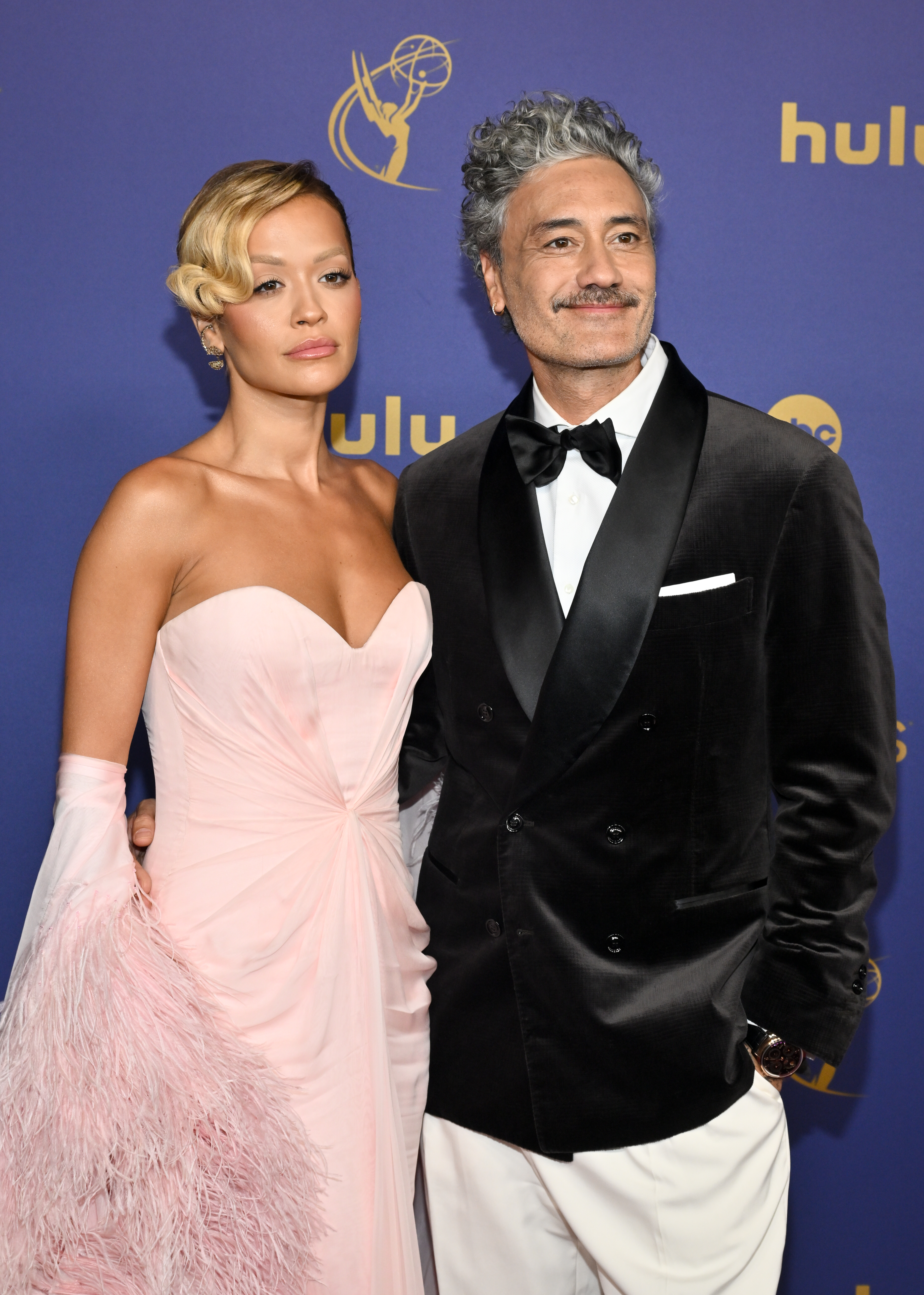 Rita Ora and Taika Waititi pose on the red carpet. Rita wears an elegant gown with feathered details, and Taika sports a classic tuxedo