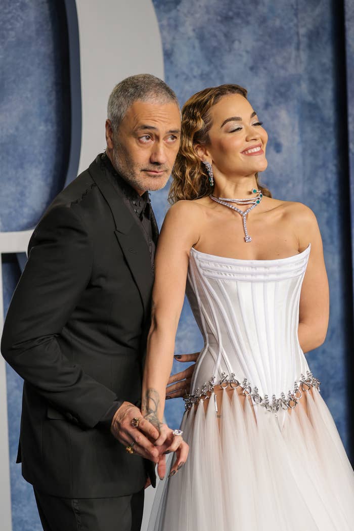 Taika Waititi in a dark suit holds hands with Rita Ora, who wears an elegant, strapless gown