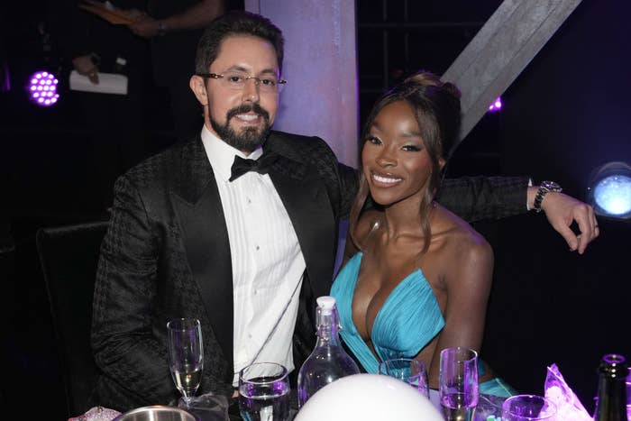 A man in a tuxedo and a woman in a low-cut blue dress are seated at a table with drinks and smiling at the camera