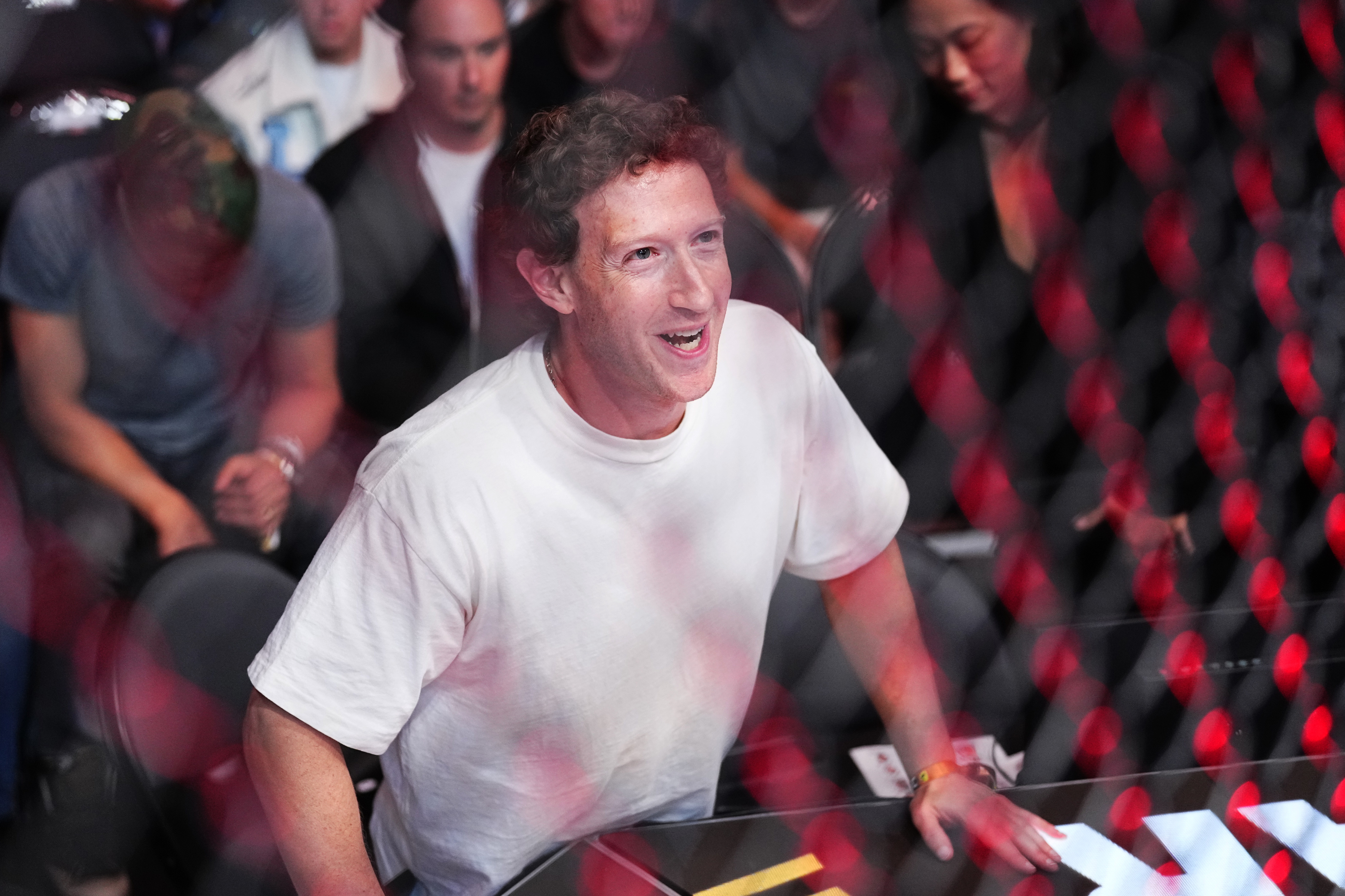 Mark Zuckerberg is seen enthusiastically cheering from behind a wire mesh fence at an event, surrounded by other attendees