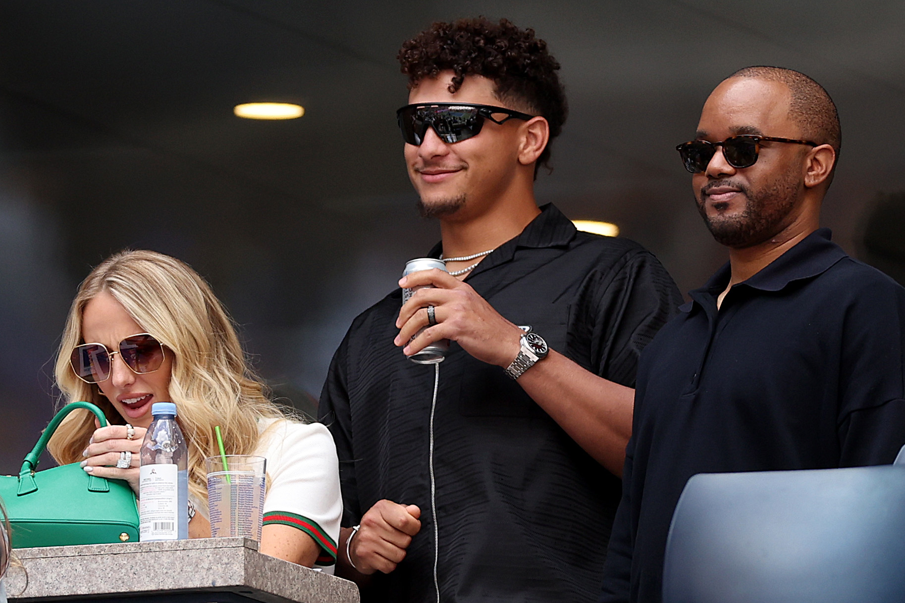 Brittany Mahomes, Patrick Mahomes, and a friend at an event, Brittany with a green handbag, Patrick holding a beverage can, all wearing sunglasses