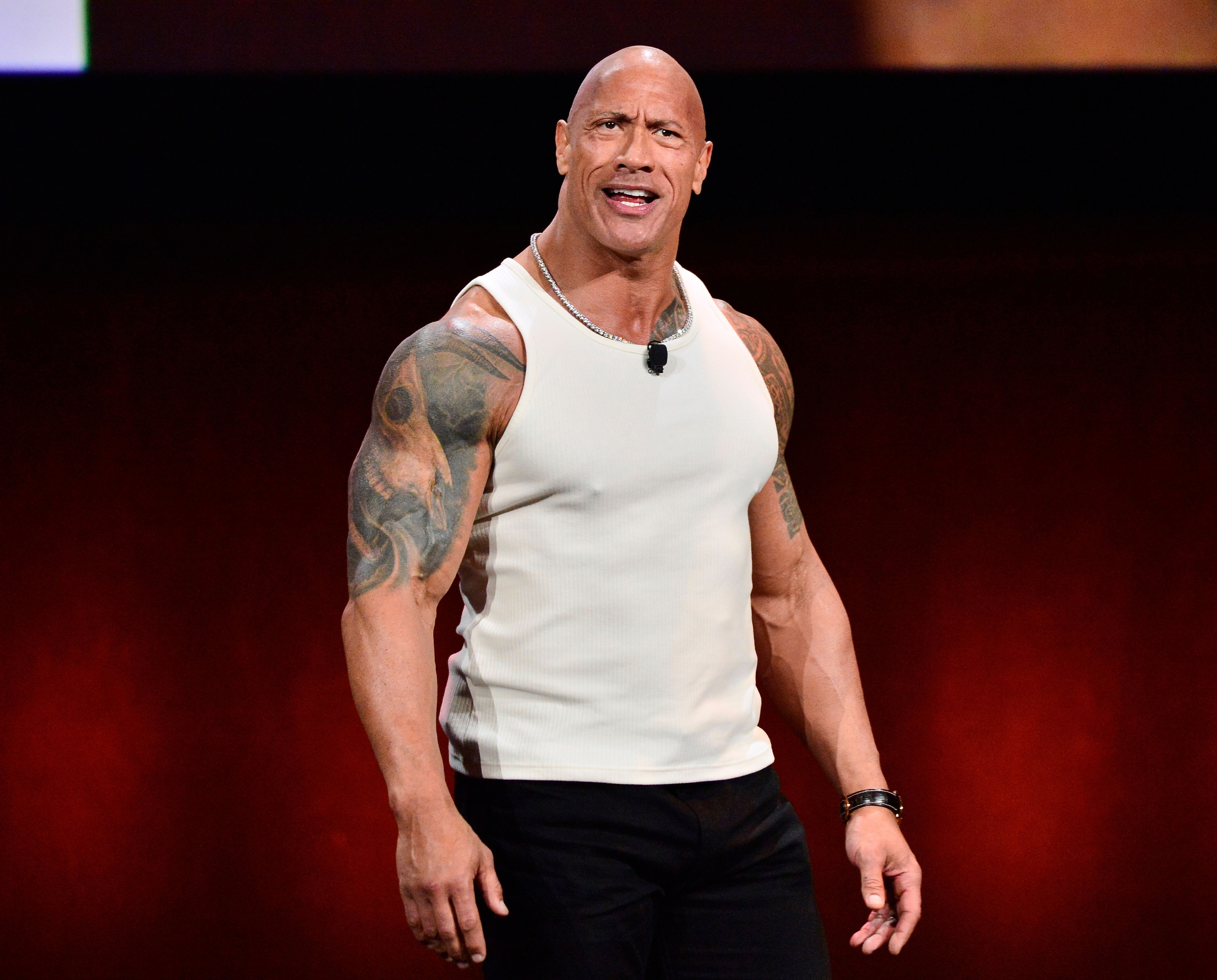 Dwayne "The Rock" Johnson wearing a sleeveless white shirt and black pants, speaking energetically onstage