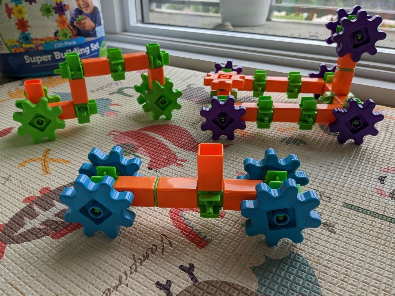 Colorful toy vehicles made from interlocking blocks with gears, displayed on a play mat near a window. Box labeled "Super Building Set" in the background