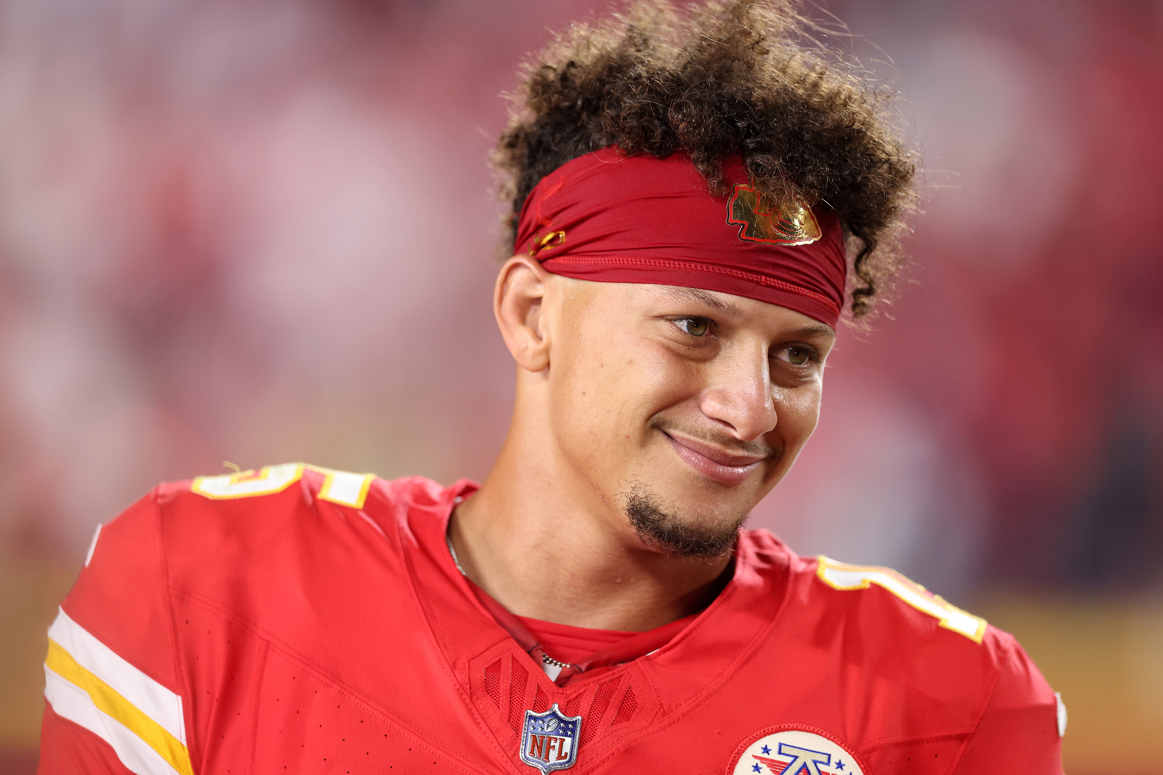Patrick Mahomes, wearing a football jersey and headband, smiles while on the field