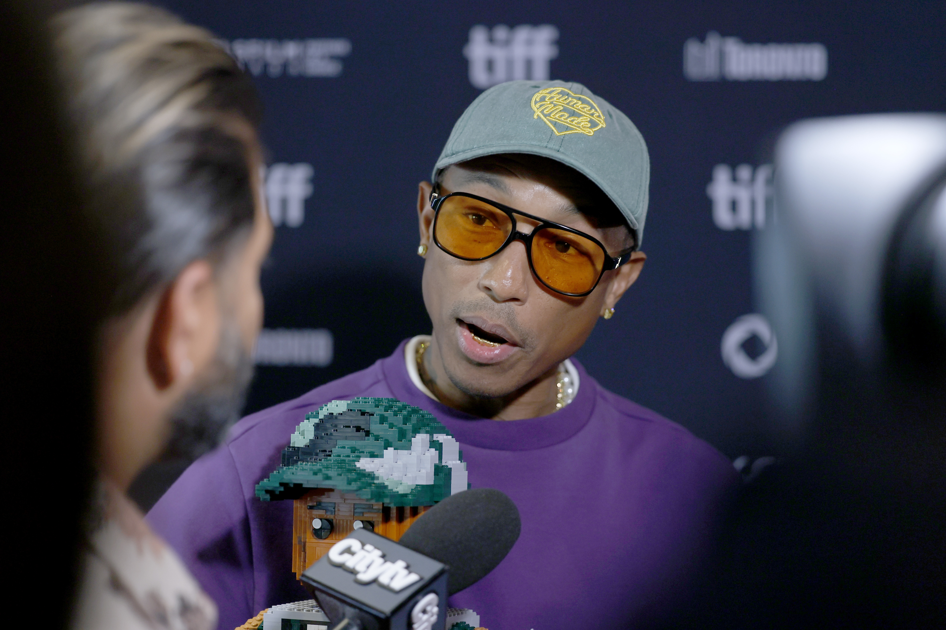 Pharrell Williams is being interviewed at a media event, wearing a green cap, sunglasses, and a purple shirt with a pixel art graphic