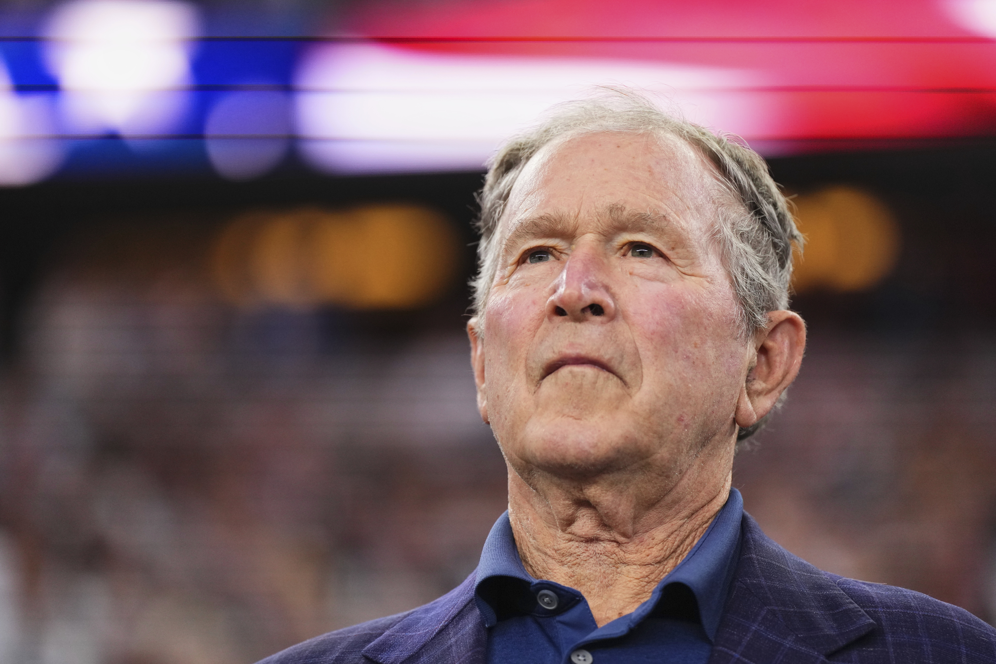 George W. Bush in a dark suit jacket and blue shirt, standing at an event with blurred lights in the background