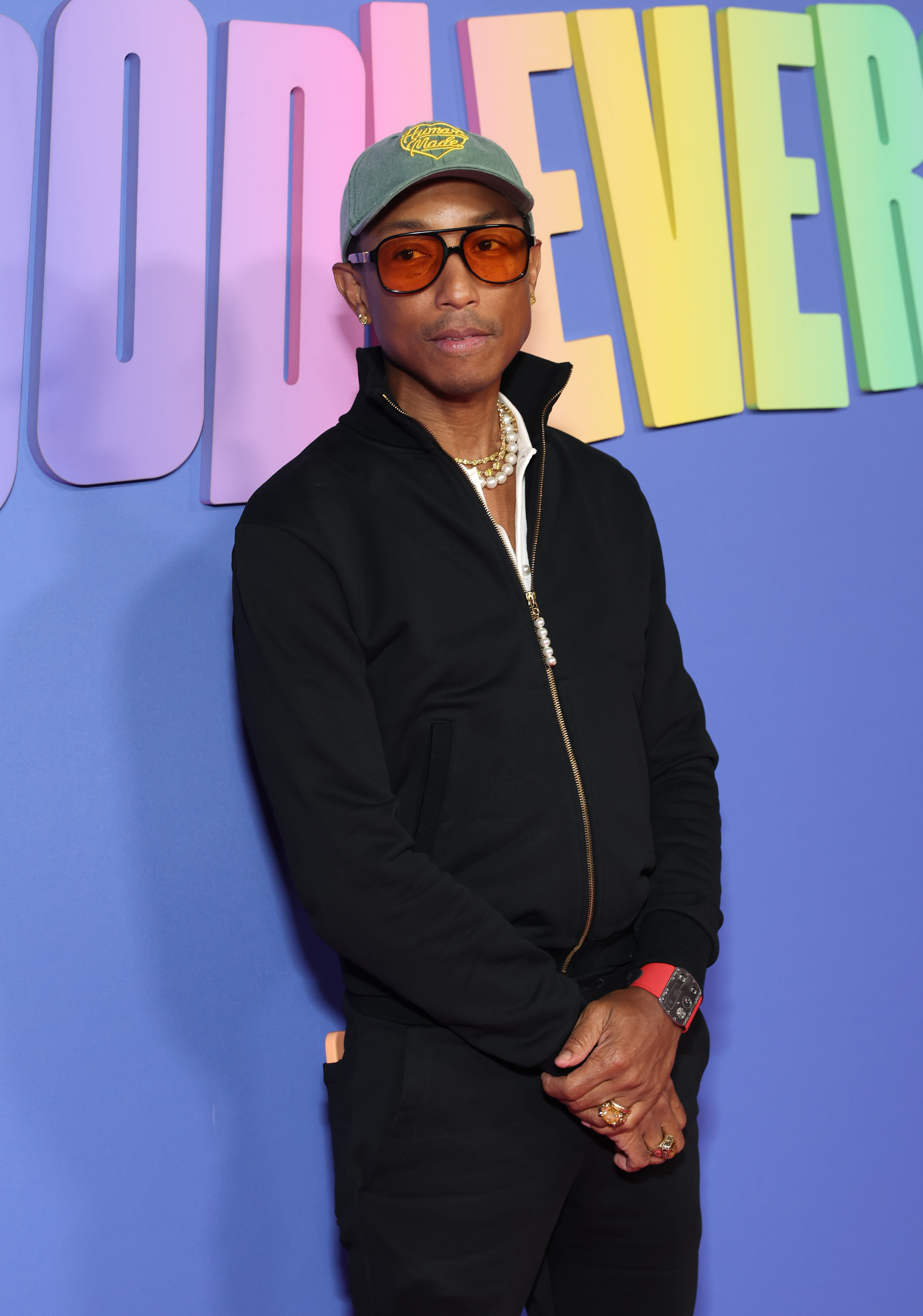 Pharrell Williams stands against a backdrop in a zip-up jacket, glasses, a hat, and multiple accessories at a celebrity event