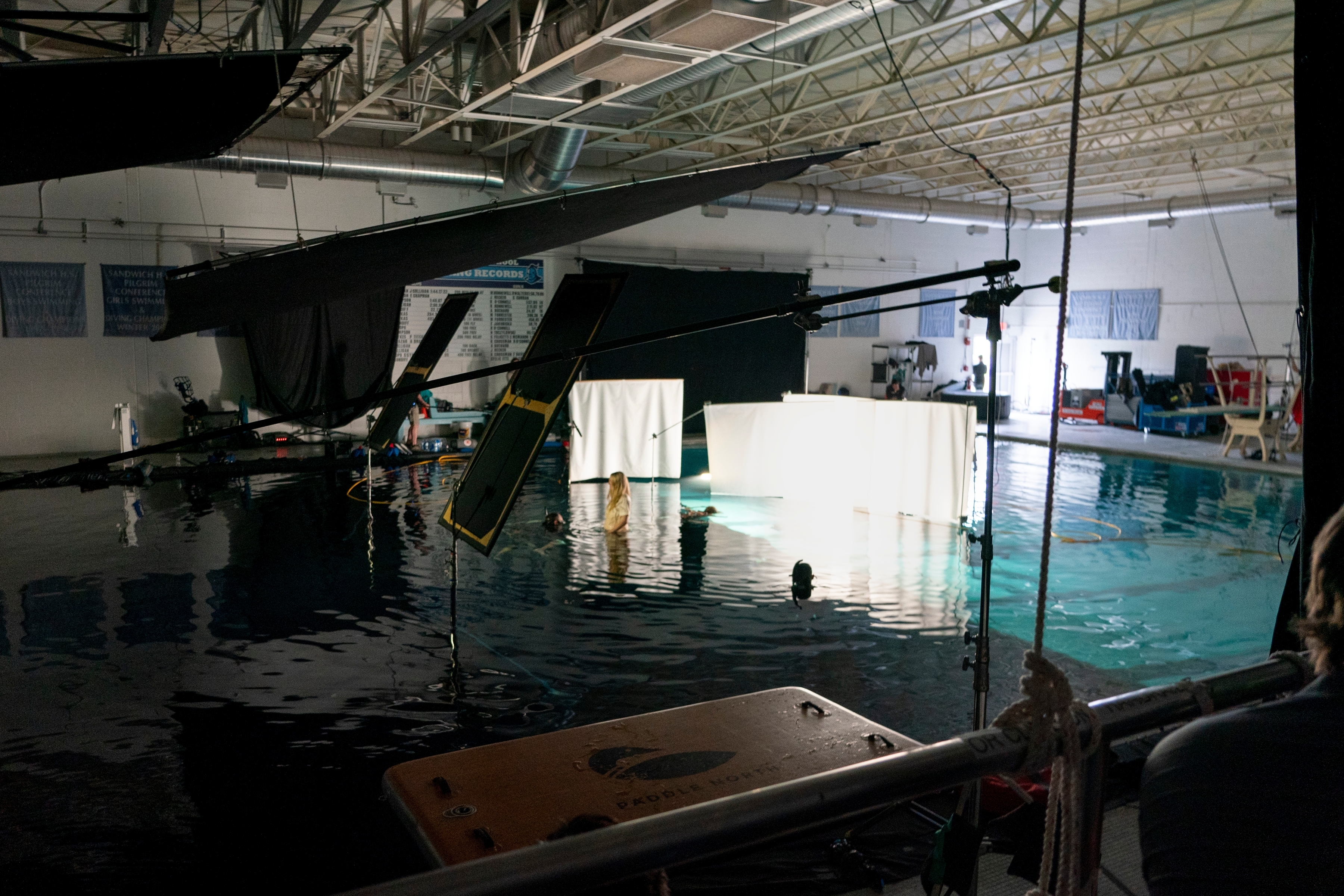 Inside a spacious film studio, crew members work around a large indoor water tank beneath heavy rigging and bright set lights, preparing for an aquatic scene