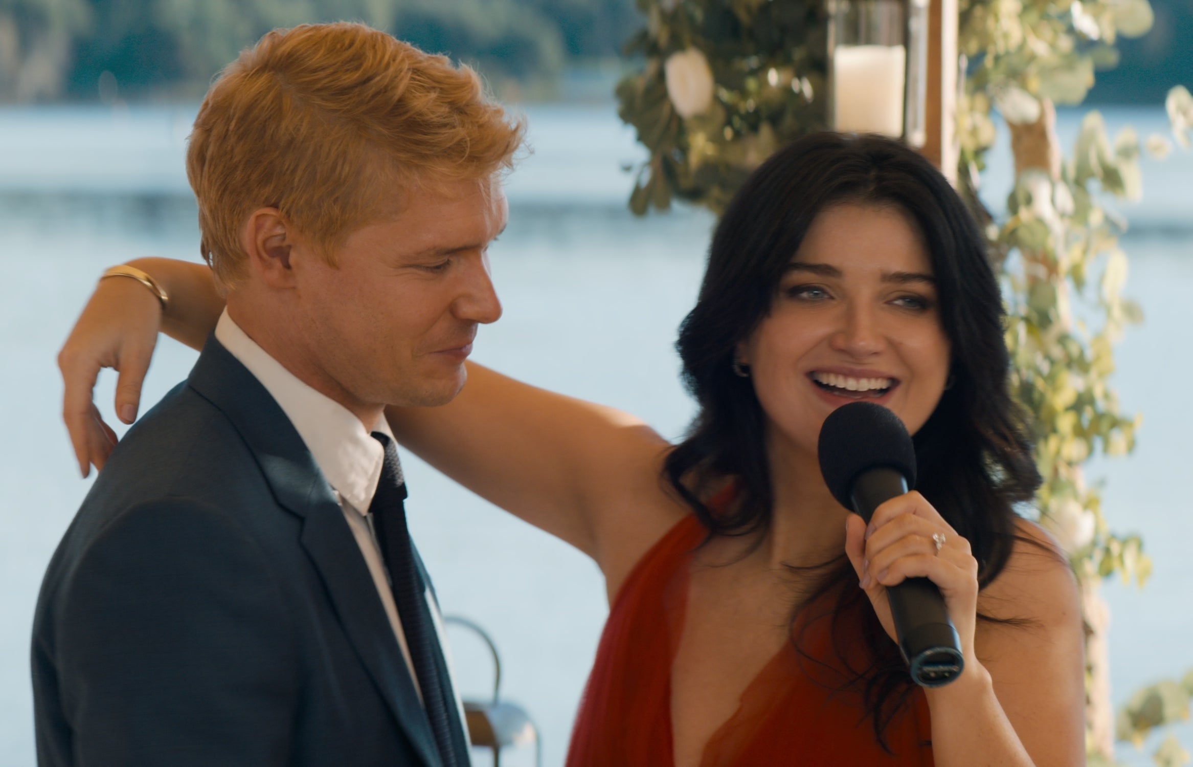 Eve Hewson, in a sleeveless dress, sings with a microphone, arm around and gazing at a man in a suit, with a scenic lake backdrop