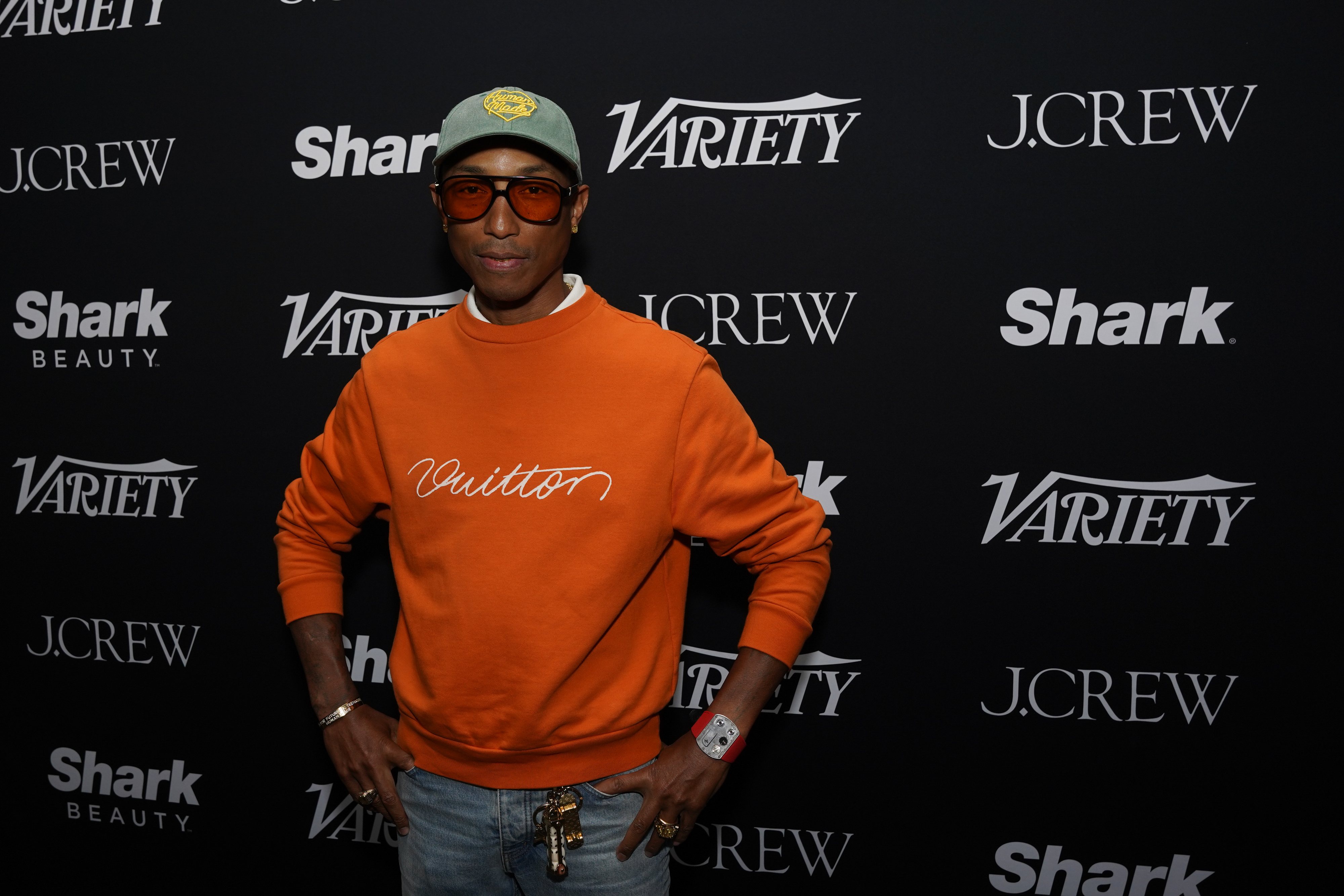 Pharrell Williams wearing a casual orange sweater with "Vuitton" text, a green cap, and red-tinted glasses at a Variety event, posing in front of a step-and-repeat backdrop