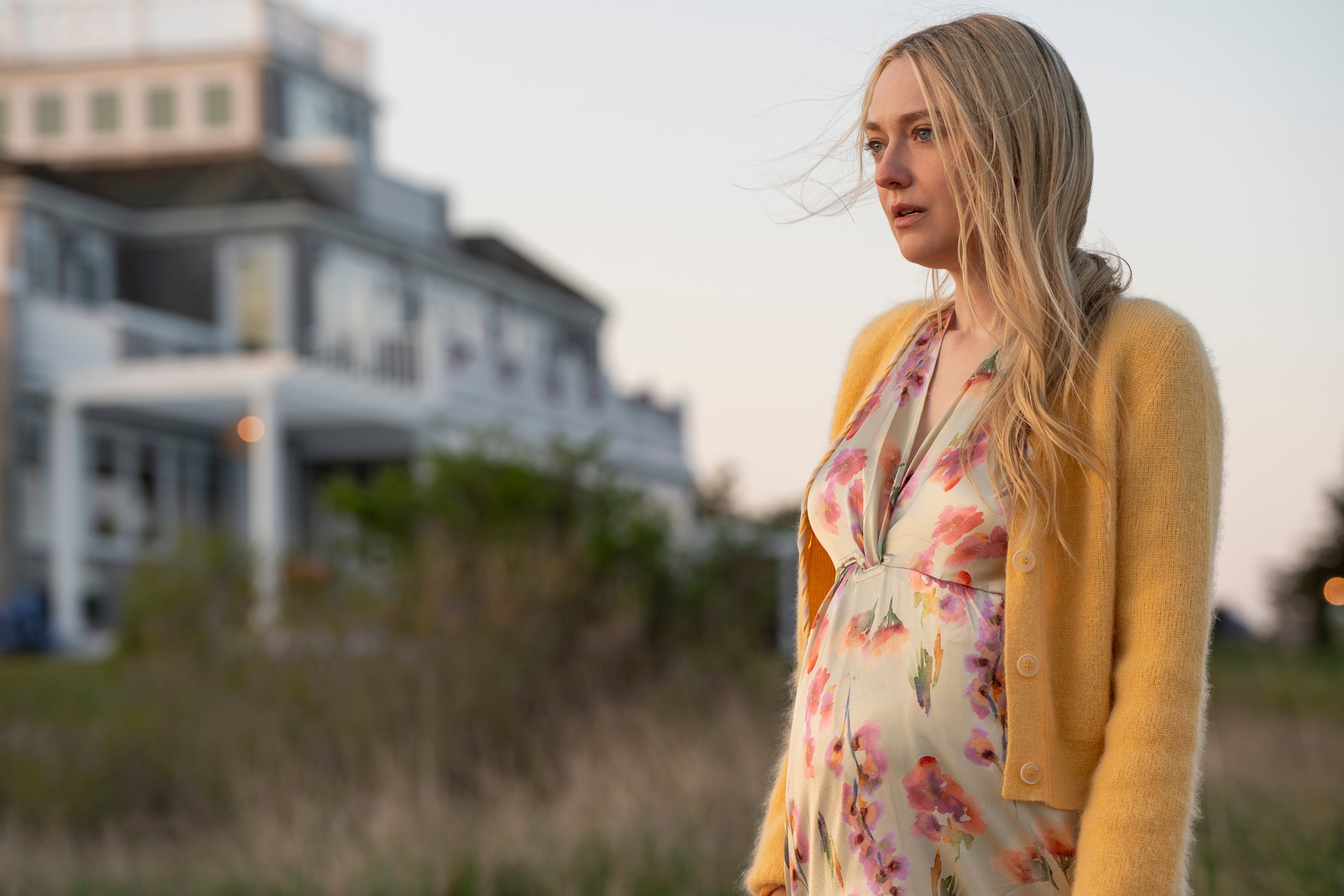 Dakota Fanning stands outdoors in front of a house, wearing a floral dress and yellow cardigan in a scene from a TV or movie