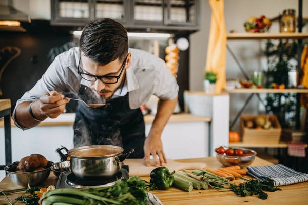 A man in a kitchen tasting soup from a ladle over a pot on the stove, with various vegetables on the counter around him
