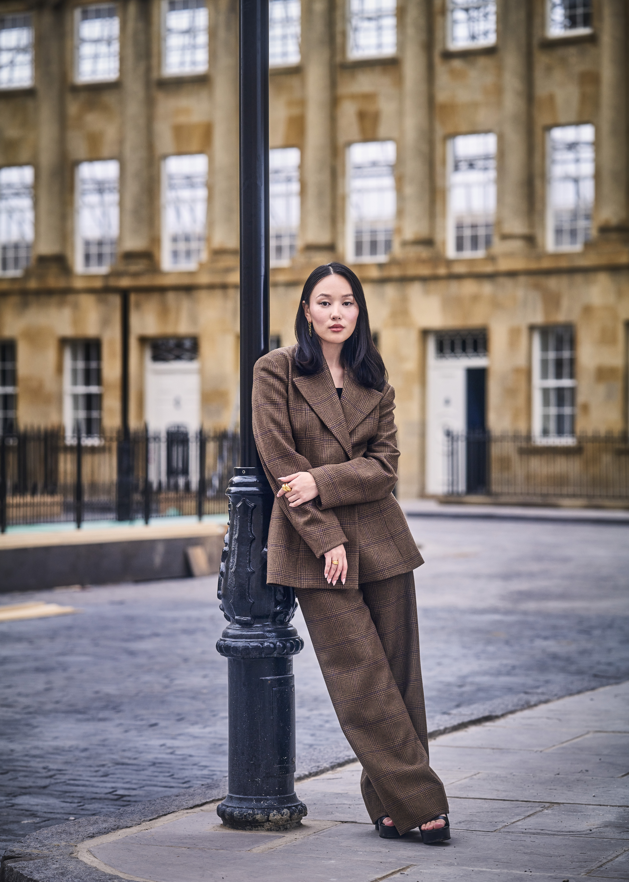 A woman wearing a tailored, brown suit leans against a lamppost in front of a historic building