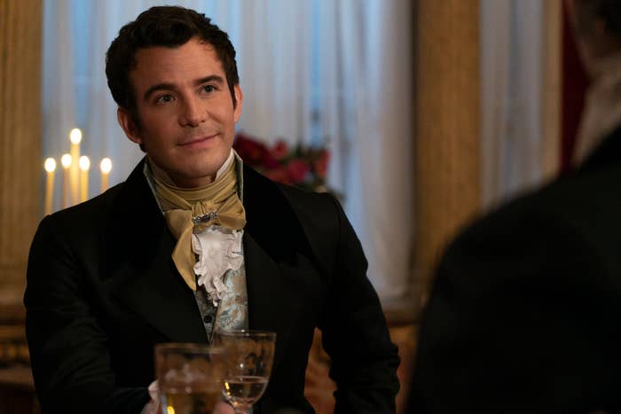 Jonathan Bailey in a period costume holds a glass, seated at a table in a lavishly decorated room with candlelight in the background