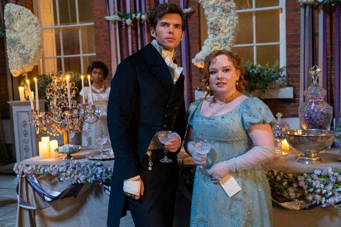 Nicola Coughlan and Luke Newton dressed in Regency-era outfits at an elegant indoor event, holding drinks and standing near a lavishly decorated table