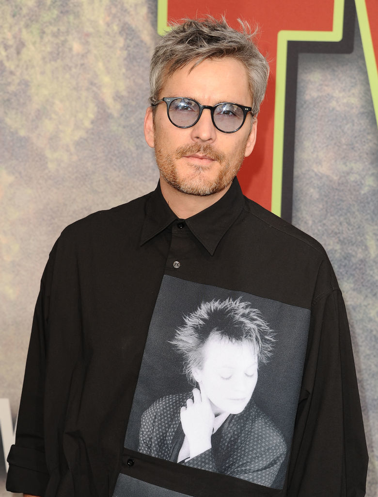 Man wearing glasses and a black shirt with a grayscale portrait graphic on it stands at a media event. Names of persons in the image unknown