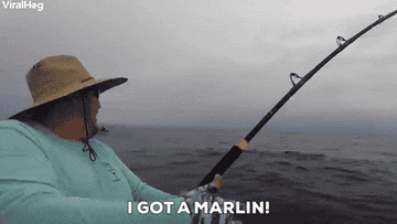 Person in a wide-brimmed hat on a boat, holding a fishing rod bent under the weight of a fish, with overlaid text reading "I GOT A MARLIN!"
