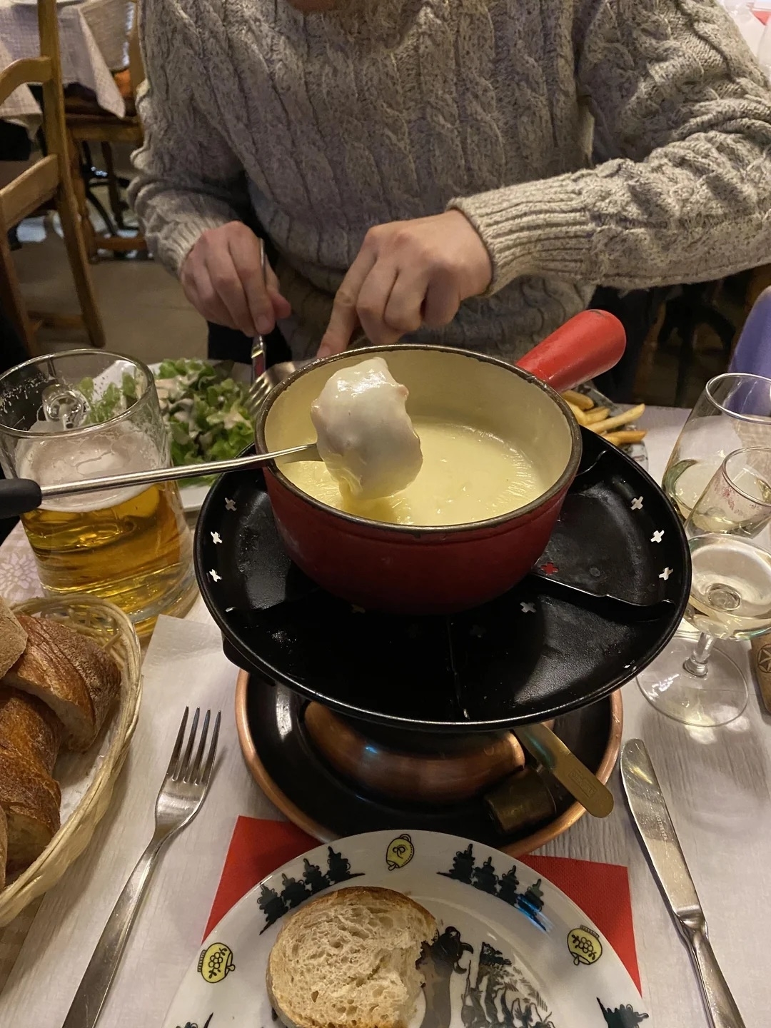 Hands dipping a piece of bread into a cheese fondue pot at a restaurant table, surrounded by bread, a beer glass, a wine glass, and cutlery