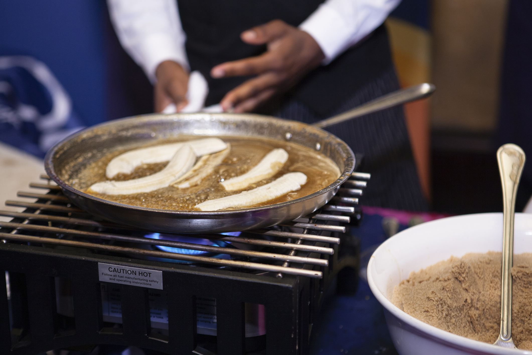 Person cooking banana slices in a pan over a flame with a bowl of brown sugar on the side