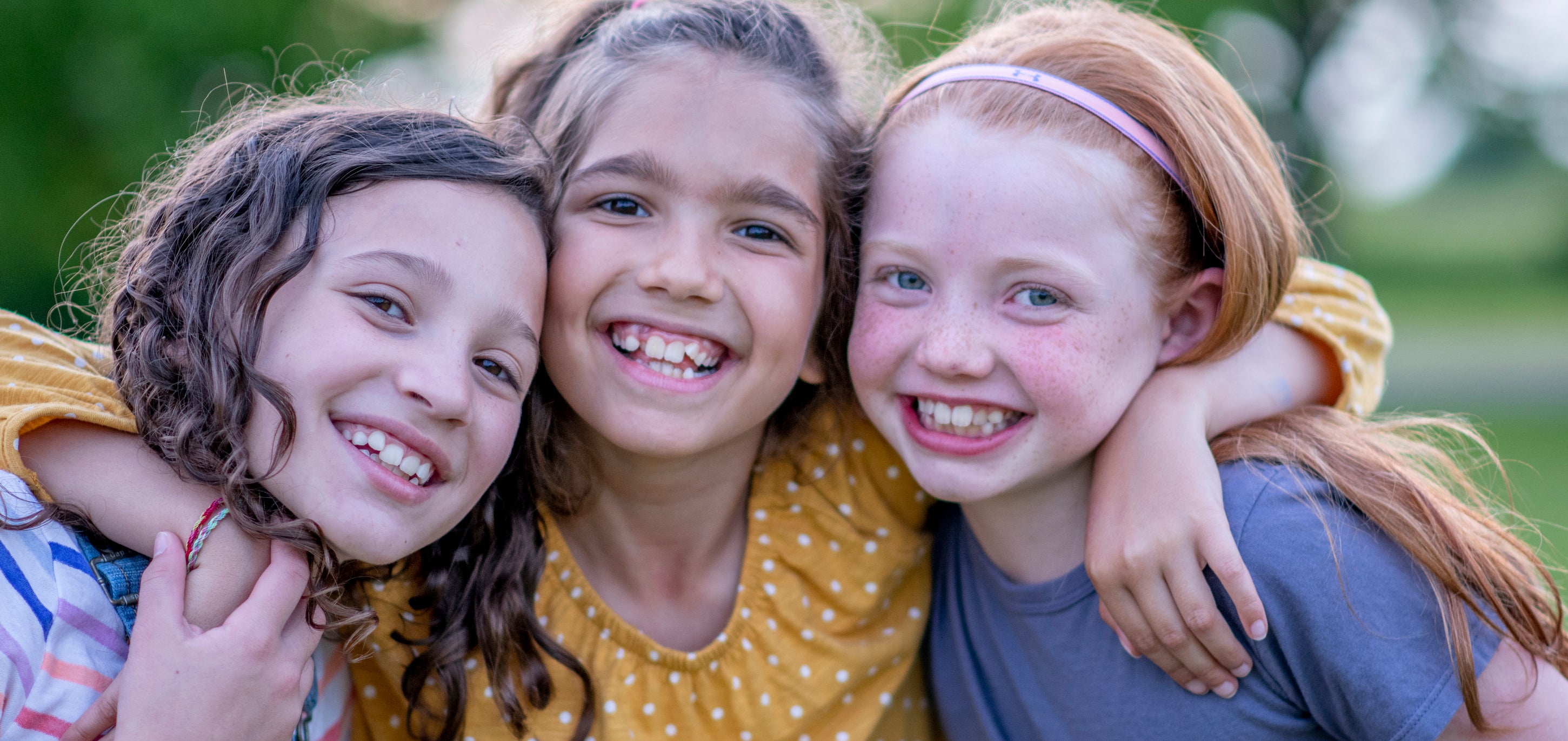 Three young girls smiling and hugging outdoors in a park