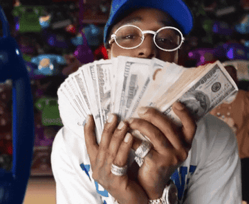 A person wearing a blue cap and glasses holds a fan of hundred-dollar bills in front of their face