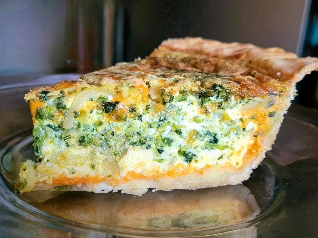 A close-up of a slice of vegetable quiche with a flaky crust, filled with spinach, onions, and cheese