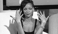 Rihanna sits on a couch with an amused expression, displaying her long fingernails with her hands raised in front of her