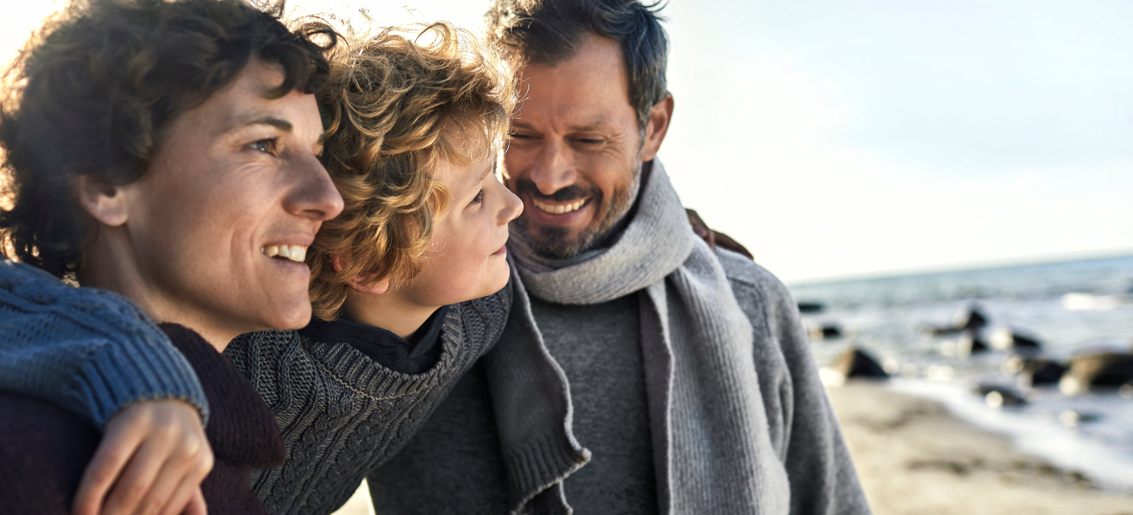 A man, woman, and child stand close together smiling on a beach, bundled in warm clothing.