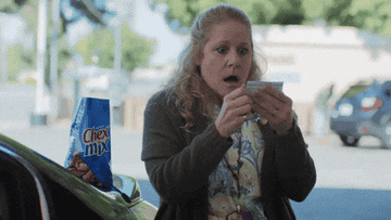 Woman looks shocked while holding a piece of paper next to a car with a Chex Mix bag on it