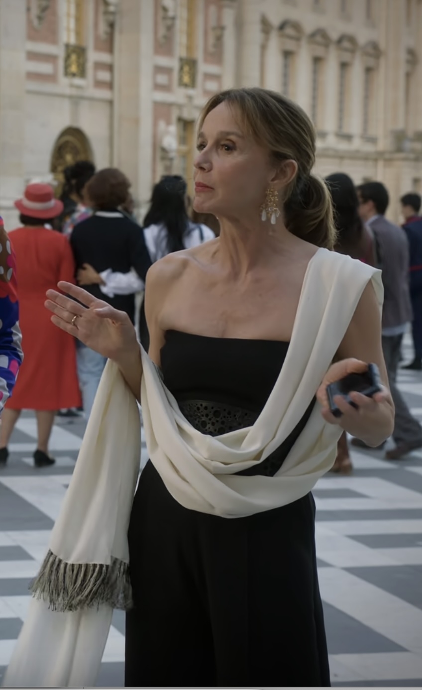 Philippine Leroy-Beaulieu  stands in an elegant strapless black dress with a white shawl draped around her, holding a phone, with a crowd and a historic building in the background