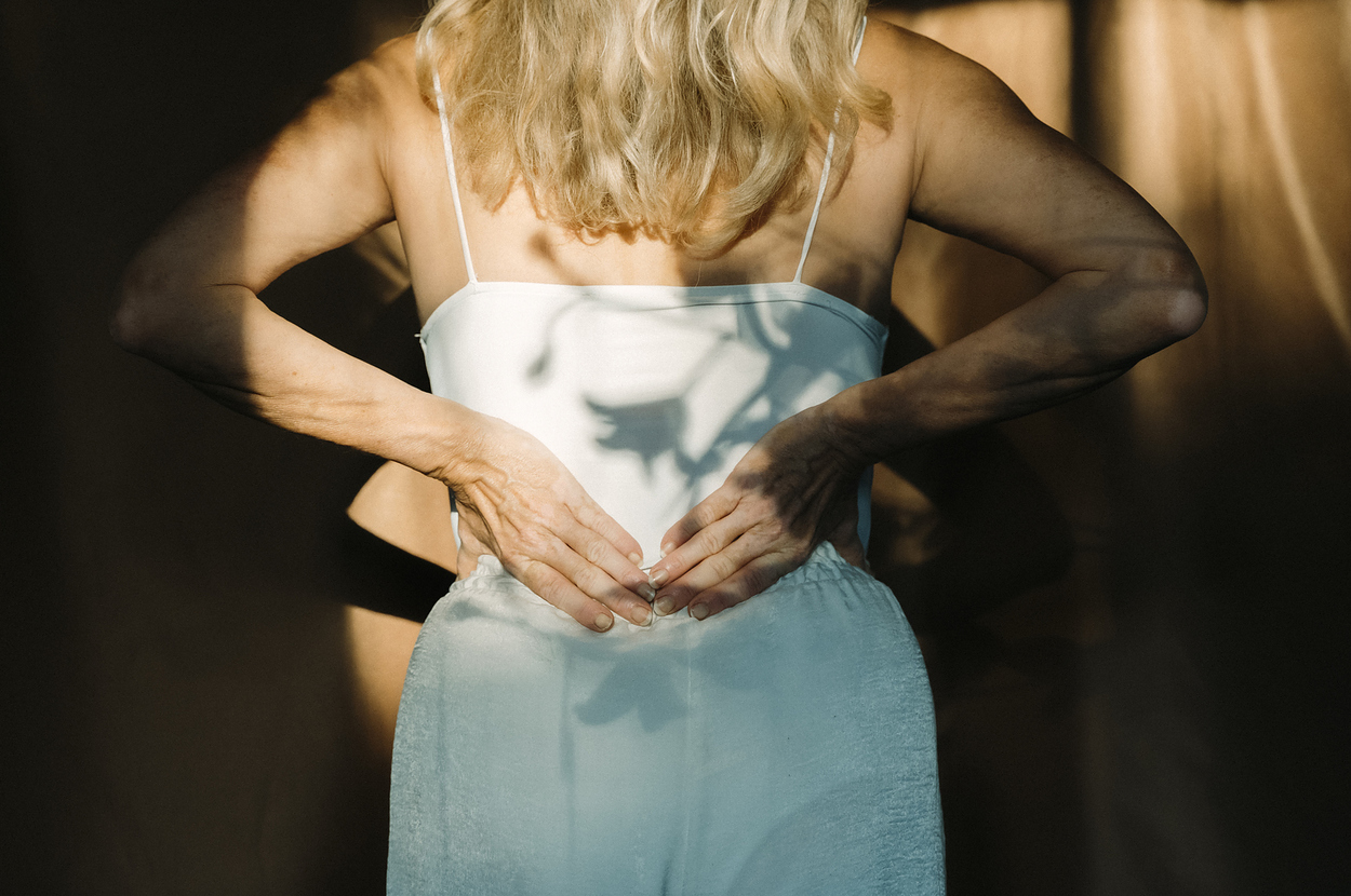 A person with long, blond hair stands facing away, adjusting the waistband of their pants. Sunlight casts shadows on their back