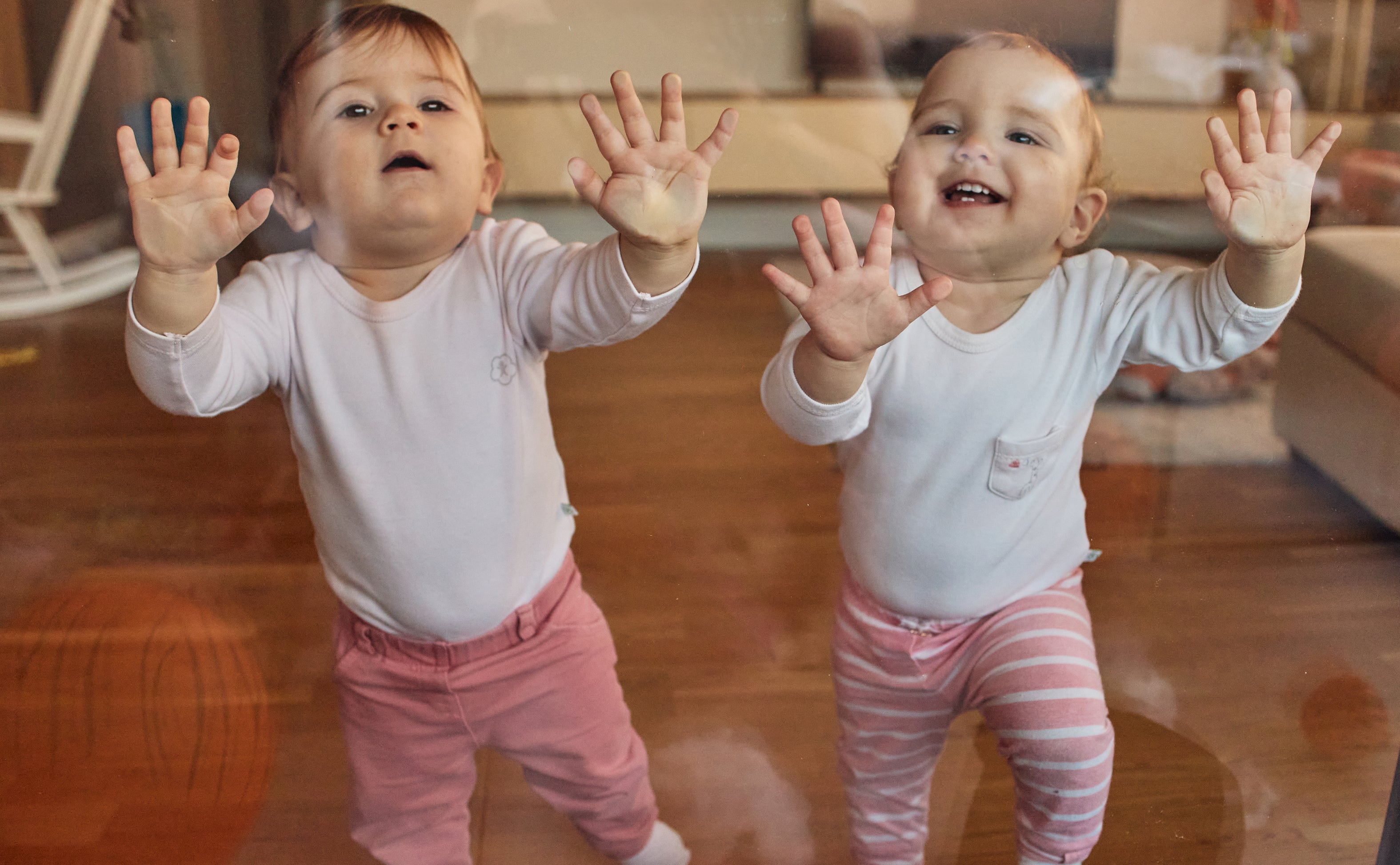 Two toddlers in matching outfits press their hands against a glass door, smiling and looking outside. The living room is visible behind them