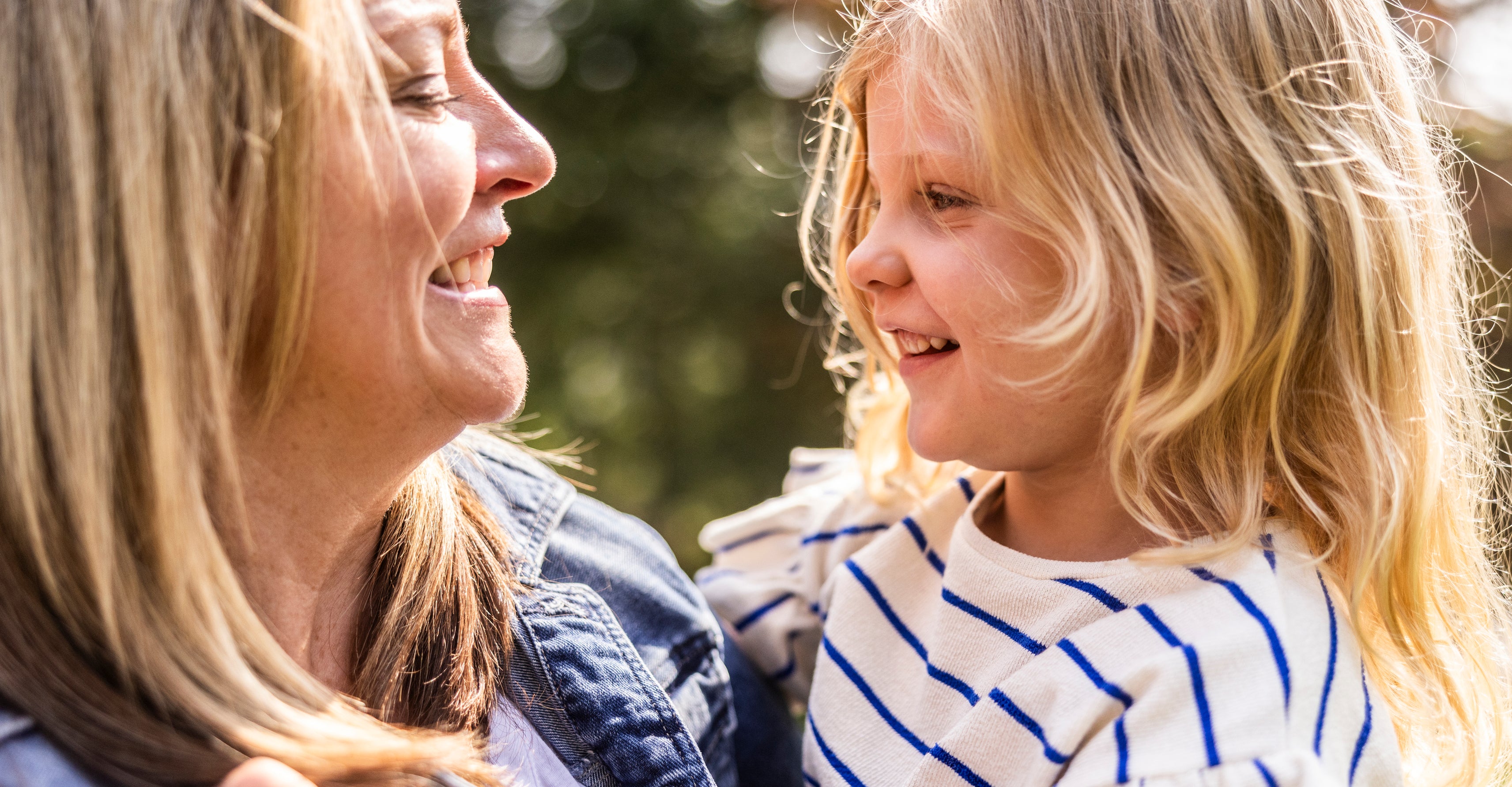 A woman and a young girl with blonde hair smile warmly at each other outdoors. The woman wears a denim jacket and the girl wears a striped top