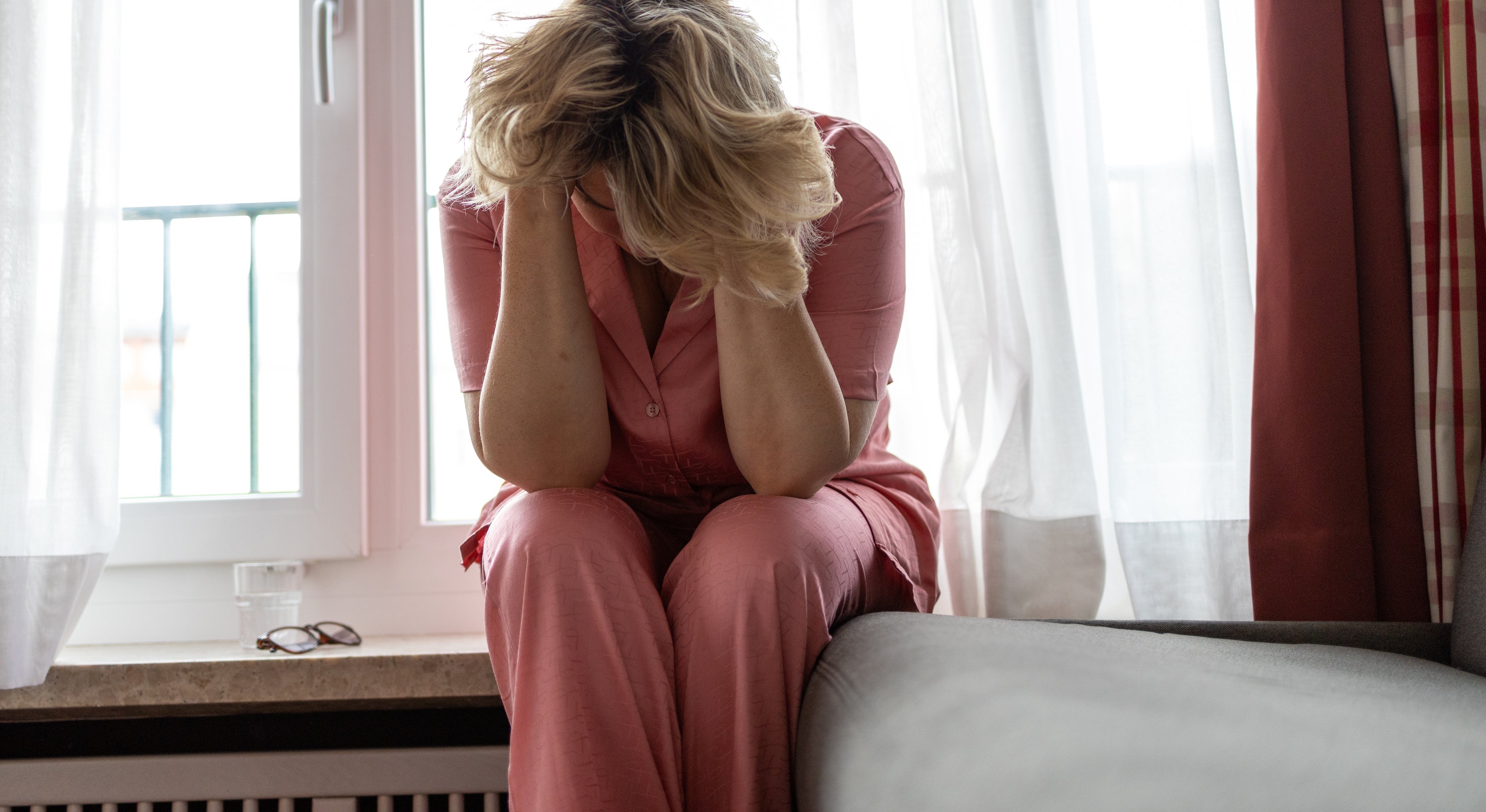 A person sits on a windowsill, head in hands, and appears distressed. Glasses lie nearby on the windowsill