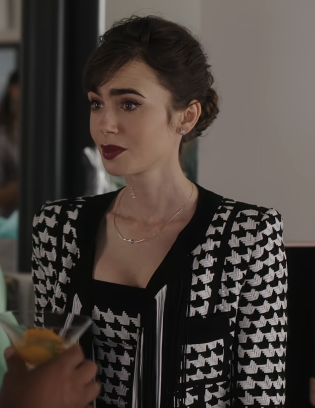 Lily Collins wearing a stylish black and white houndstooth-patterned jacket, standing indoors and looking slightly to the side