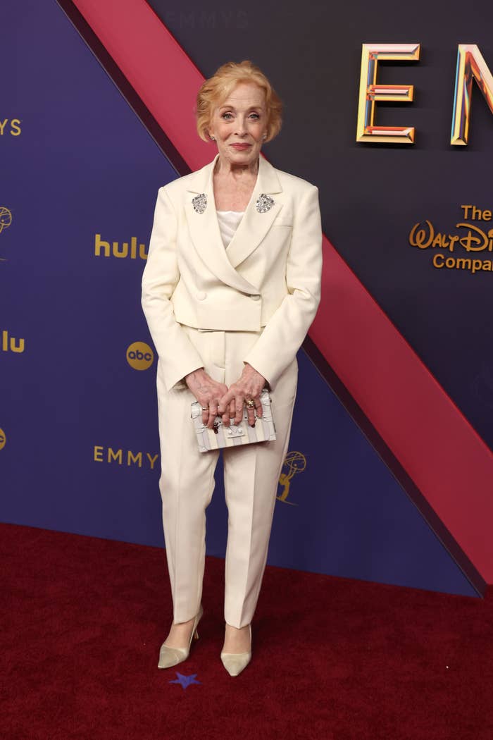 Holland Taylor standing on the red carpet wearing a white tailored suit and holding a silver clutch at an Emmy Awards event