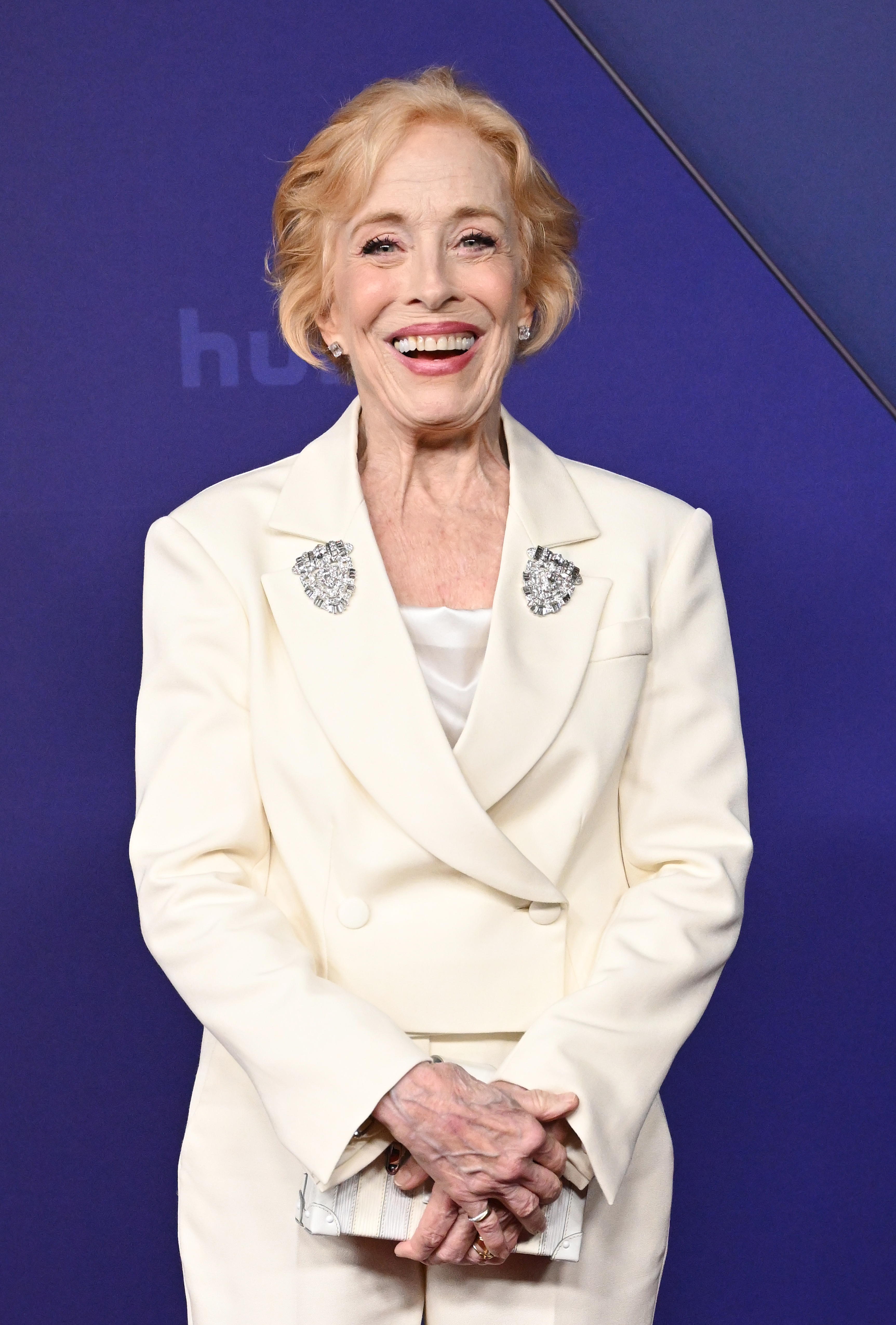 Holland Taylor smiles on a red carpet wearing a stylish white suit with decorative brooches. She holds a clutch bag