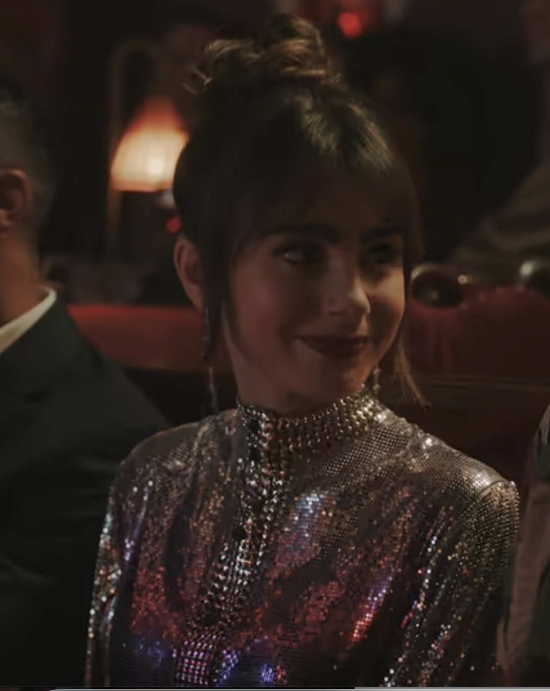 Lily Collins in a sequined dress, with her hair in a high bun, smiles while sitting in a dimly lit room