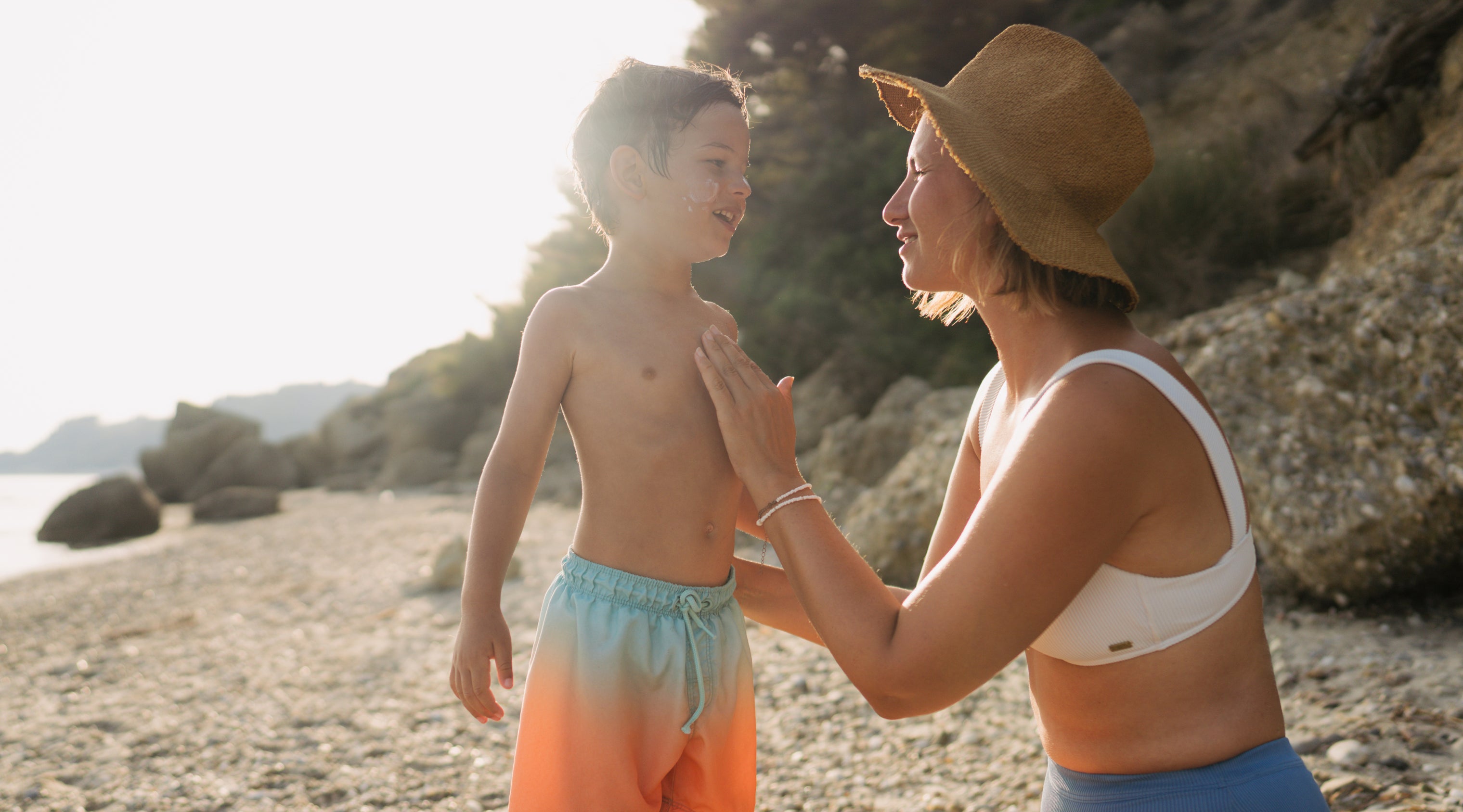 A woman wearing a straw hat and a bikini kneels on a sandy beach, smiling at a young boy in swim trunks standing in front of her