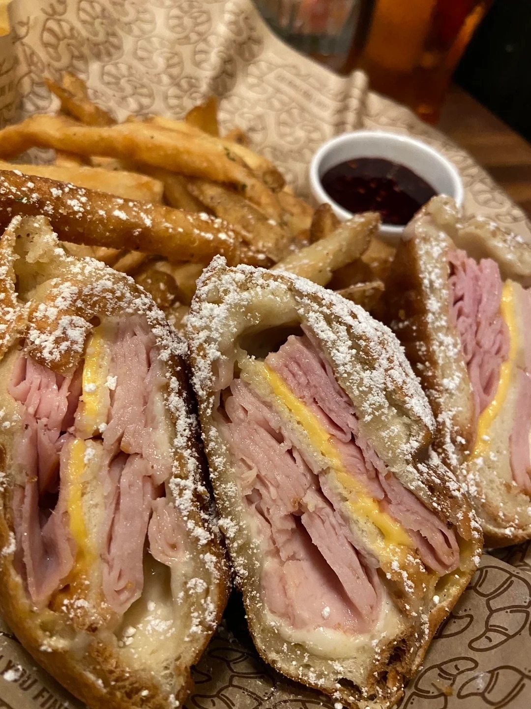 Close-up of a Monte Cristo sandwich, sliced in half, with ham, cheese, and powdered sugar, accompanied by a side of fries and jam in the background
