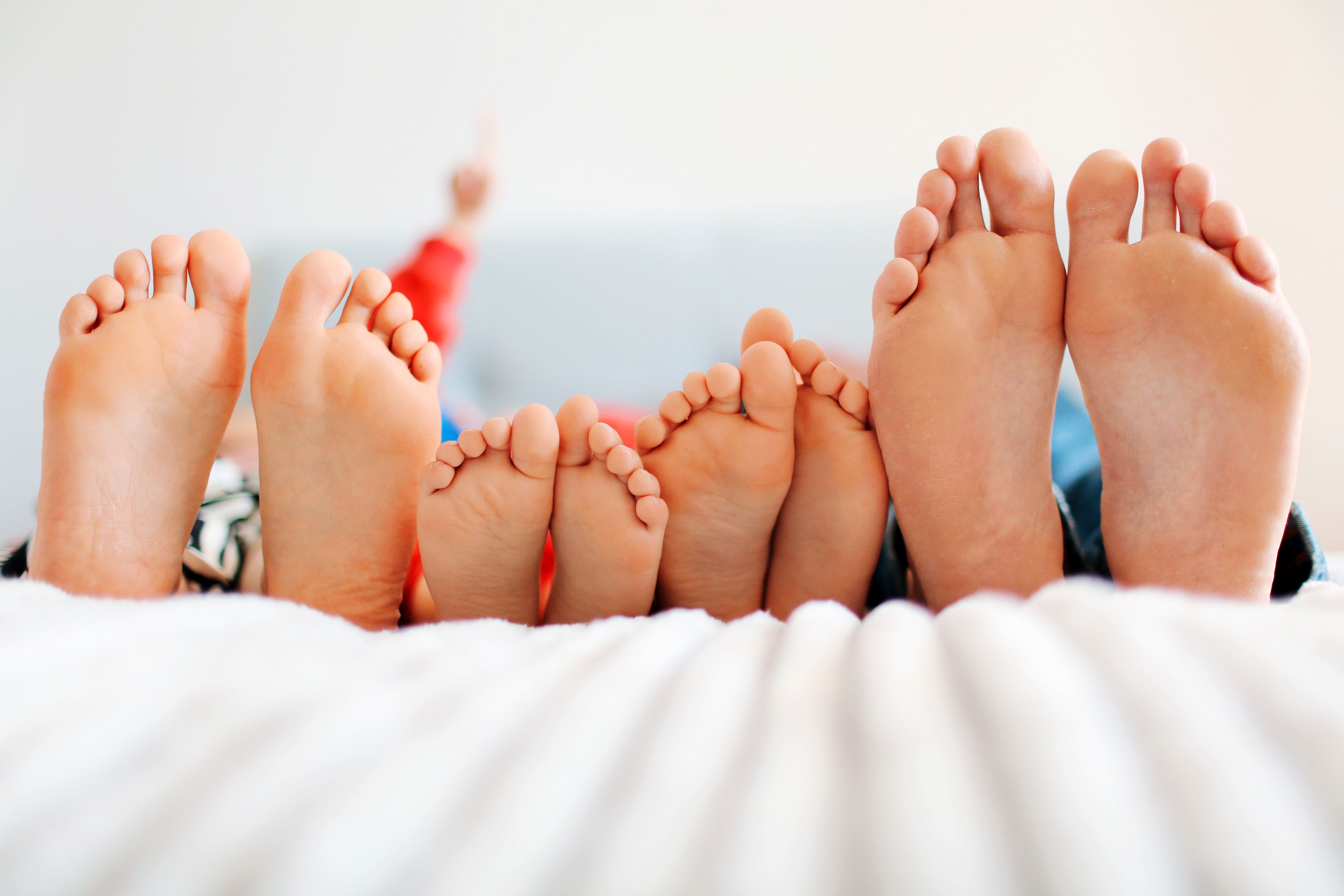 Five pairs of feet, of various sizes, are lying on a bed, pointing upwards. A person in the background is blurry, extending a finger.