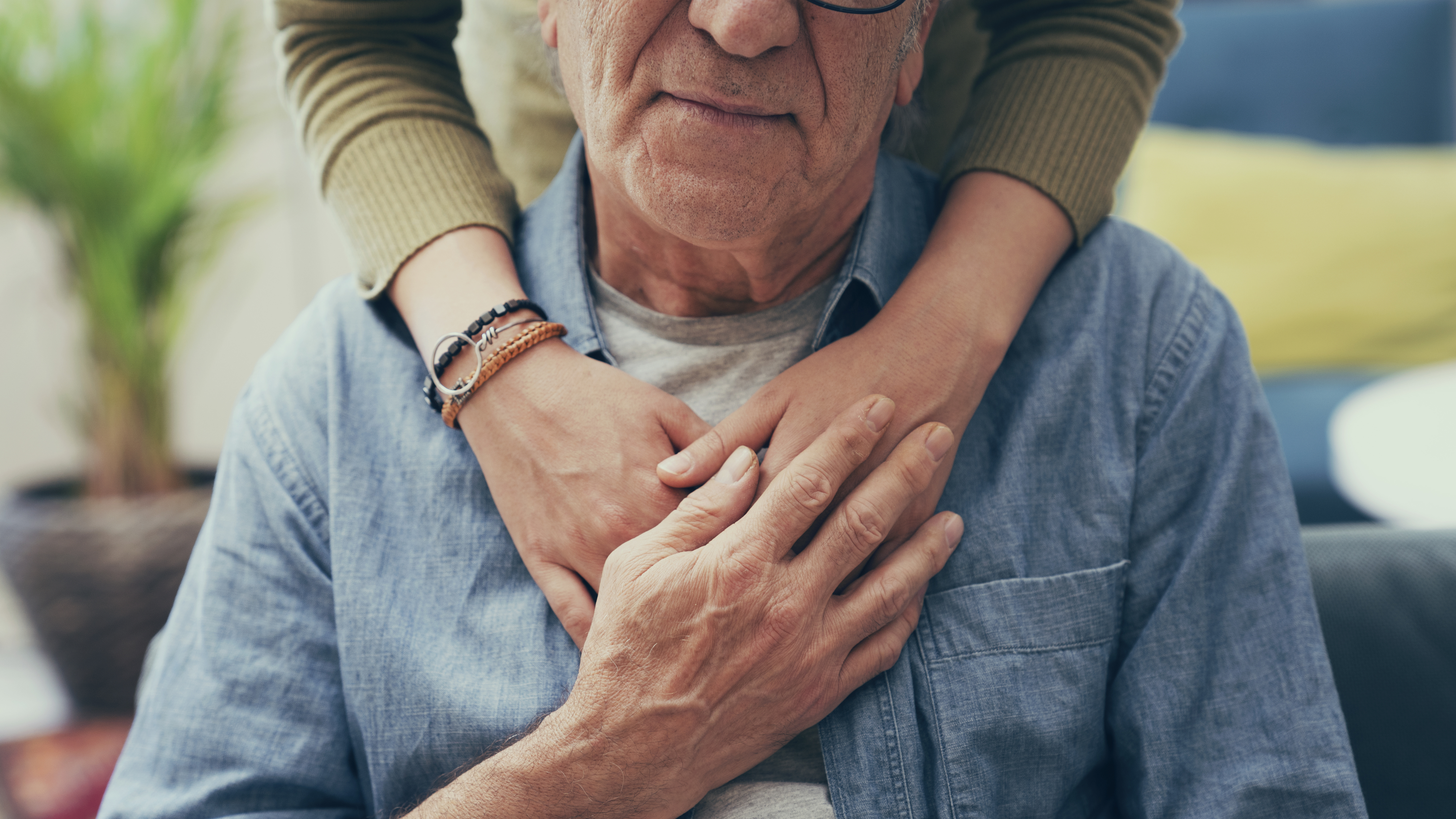 An older man wearing glasses and a denim shirt is sitting down. A younger person in a green sweater stands behind him and gently embraces him