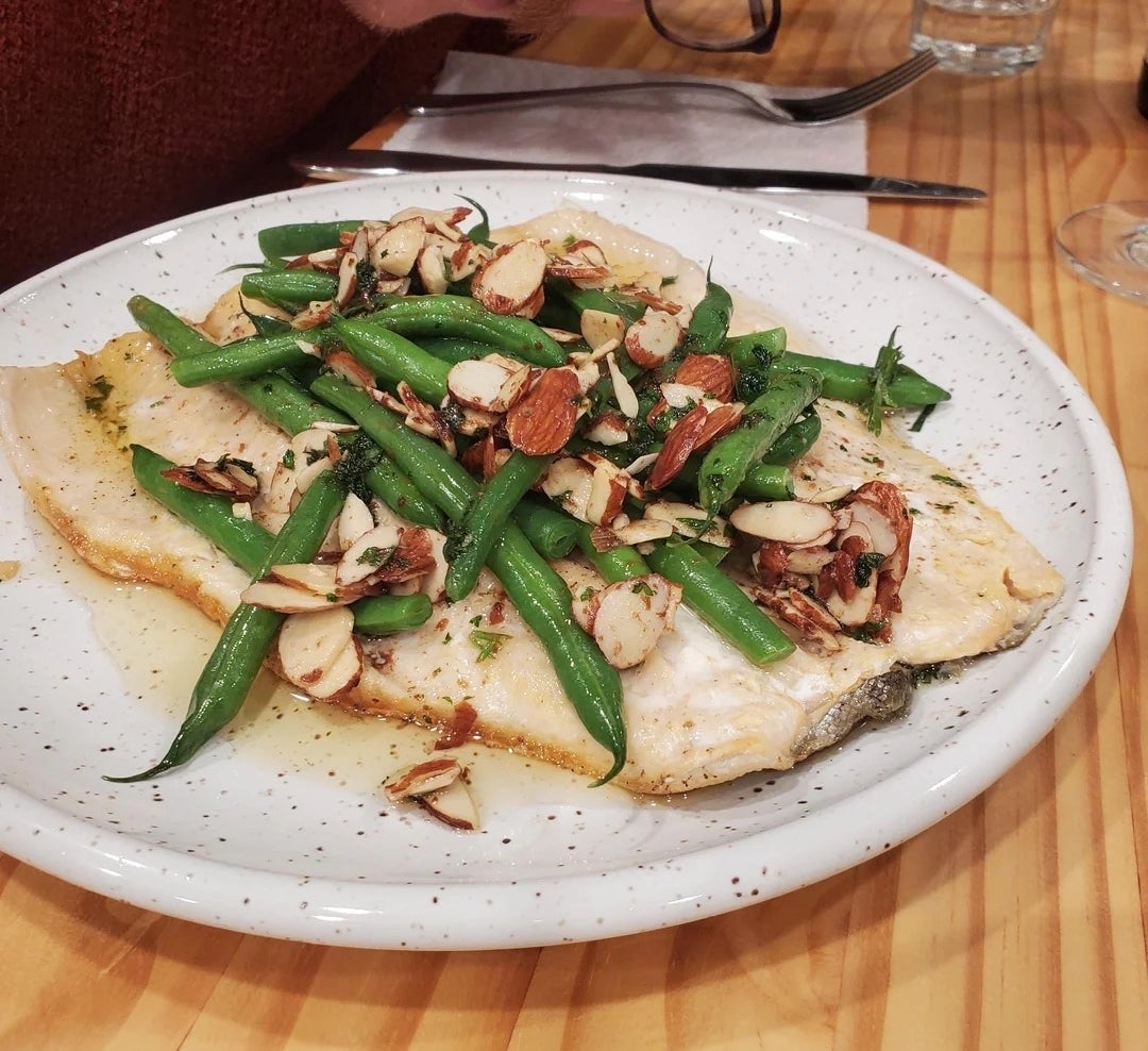 Person is leaning over a plate of fish fillet topped with green beans and sliced almonds on a wooden table