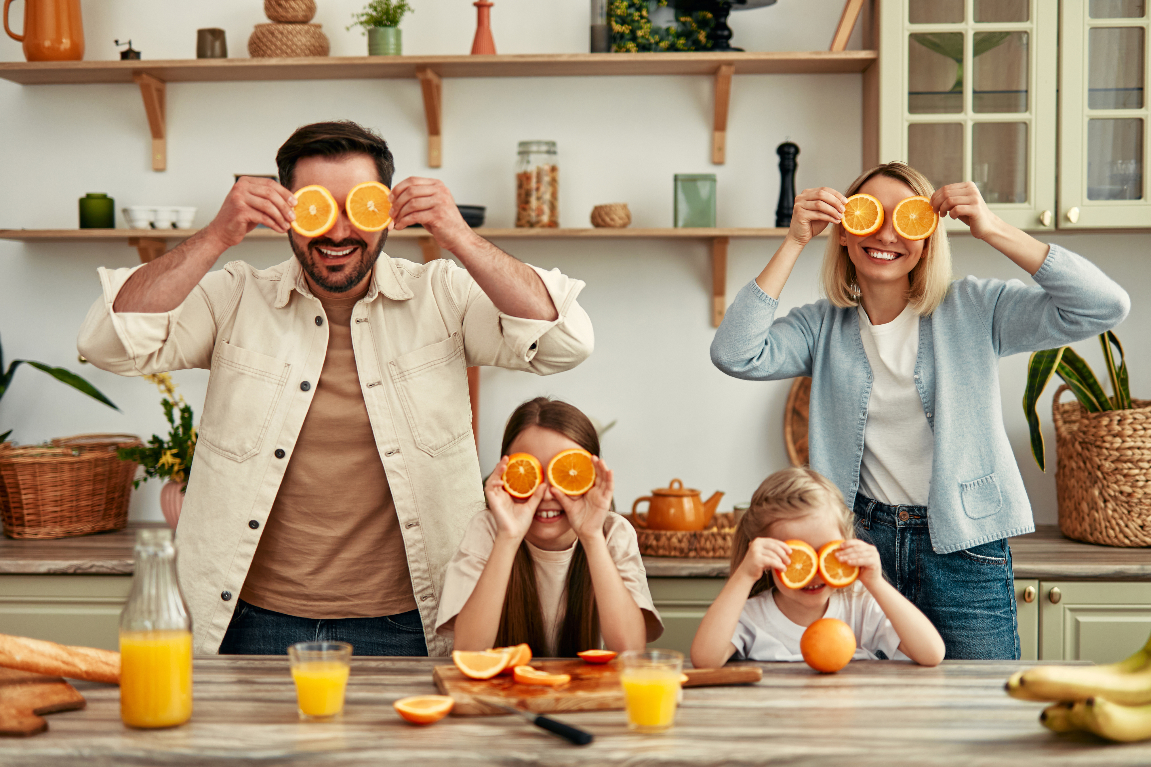 A family of four in a kitchen, holding orange slices over their eyes and smiling. The countertops are filled with various fruits and kitchen items
