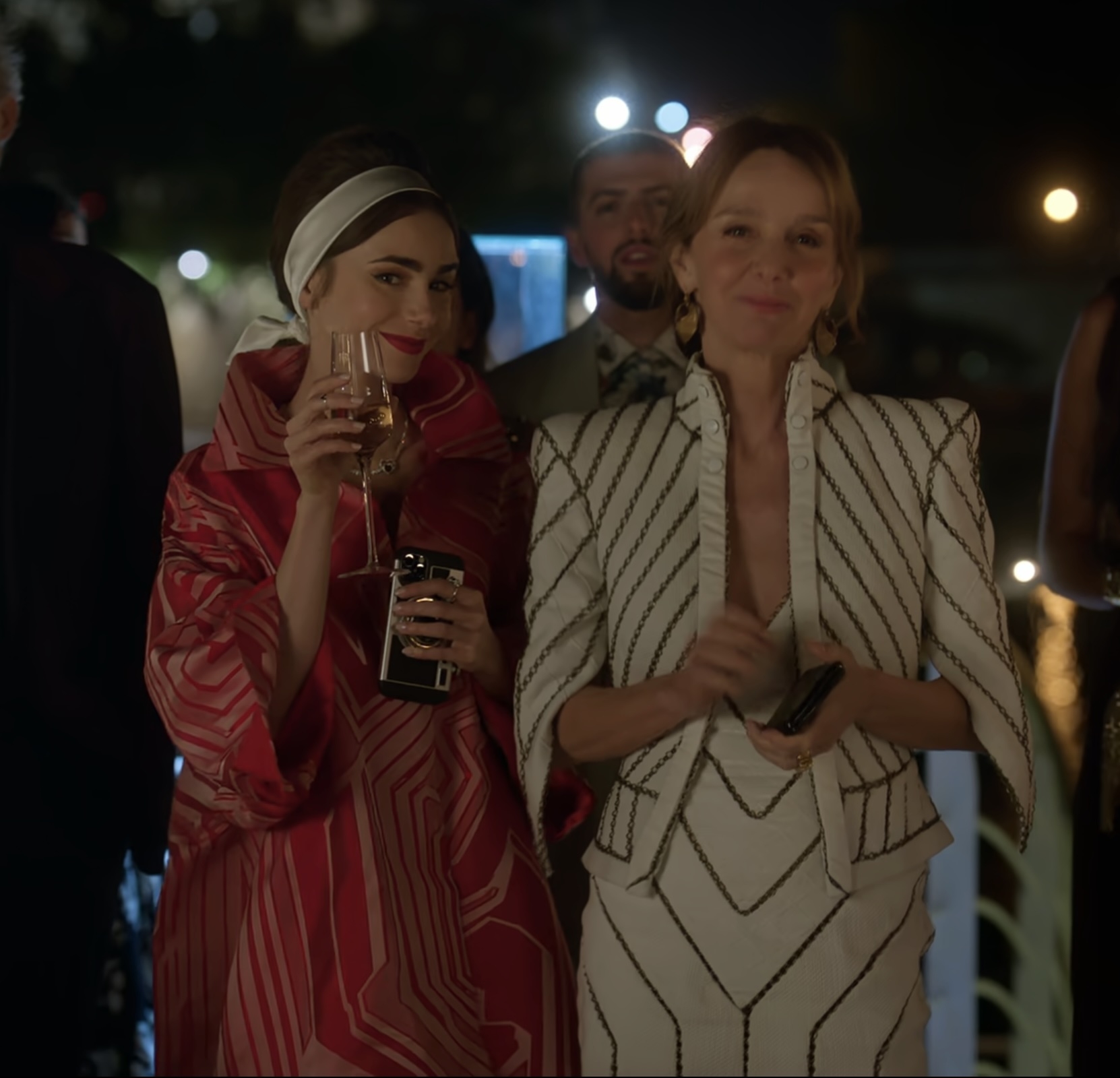 Lily Collins and Philippine Leroy-Beaulieu smile at an outdoor evening event. Lily wears a flowing, patterned outfit with a headband, while Philippine wears a structured, striped jacket