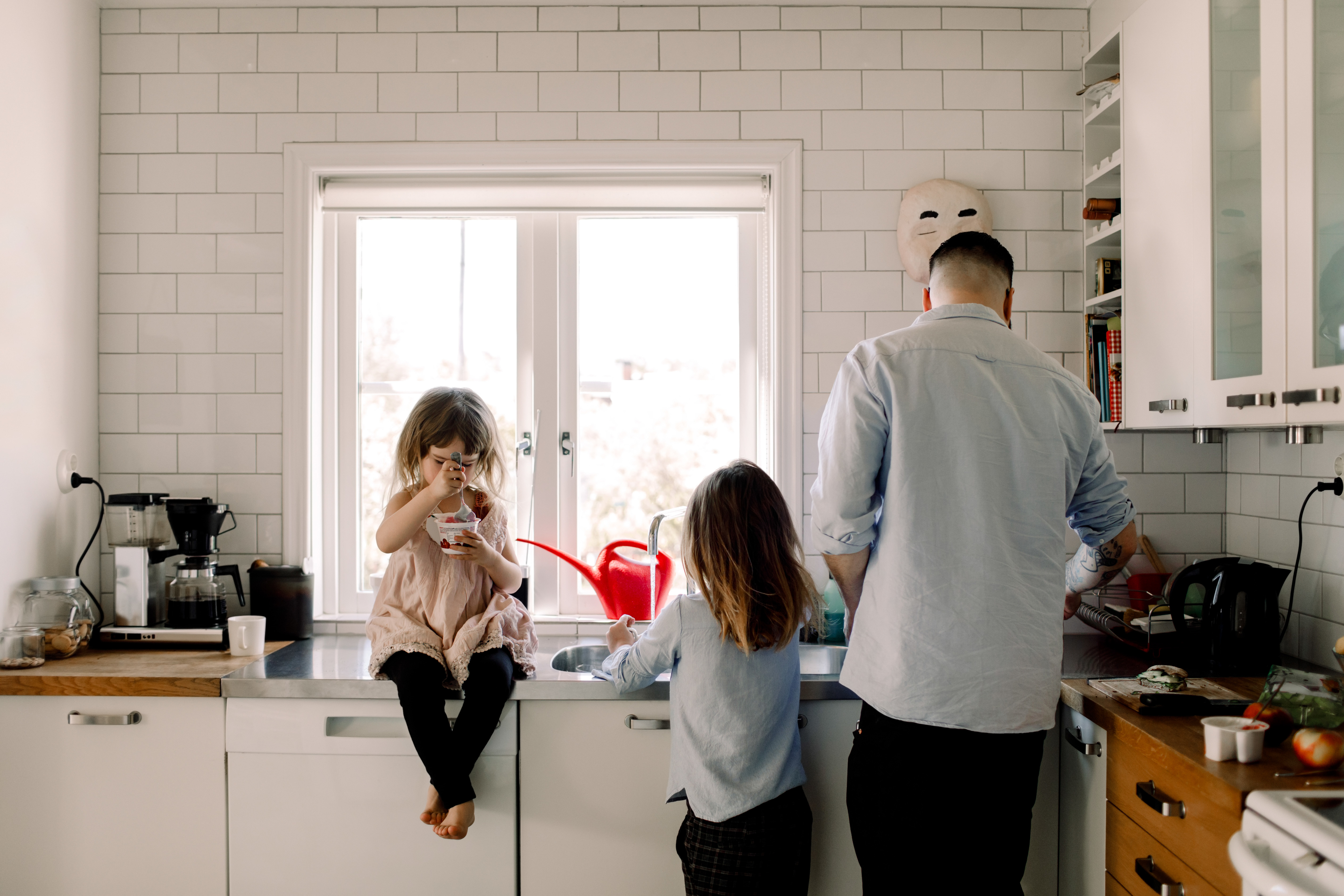 Two children sit on a kitchen counter while a man prepares food. A white mask hangs on the wall