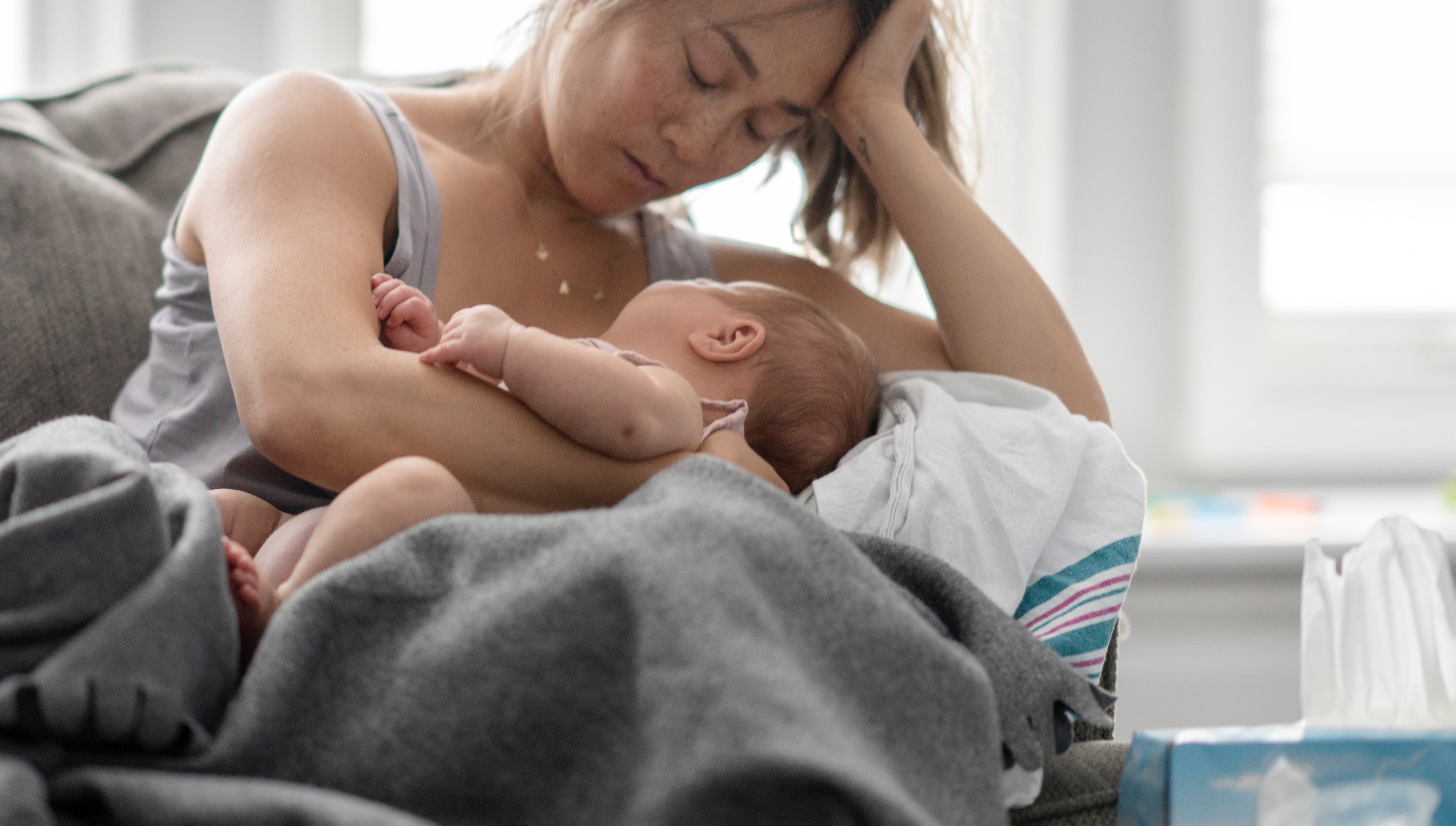 Mother holding a sleeping baby while seated on a couch, looking tired with her head resting on one hand and surrounded by a blanket and tissues