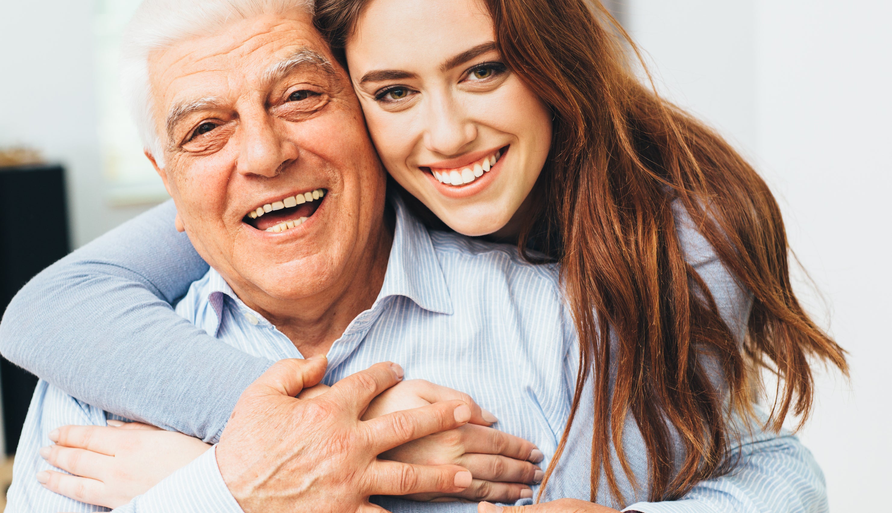 An elderly man and a young woman, both smiling, embrace warmly in a well-lit room. The man wears a light-colored shirt, and the woman has long hair